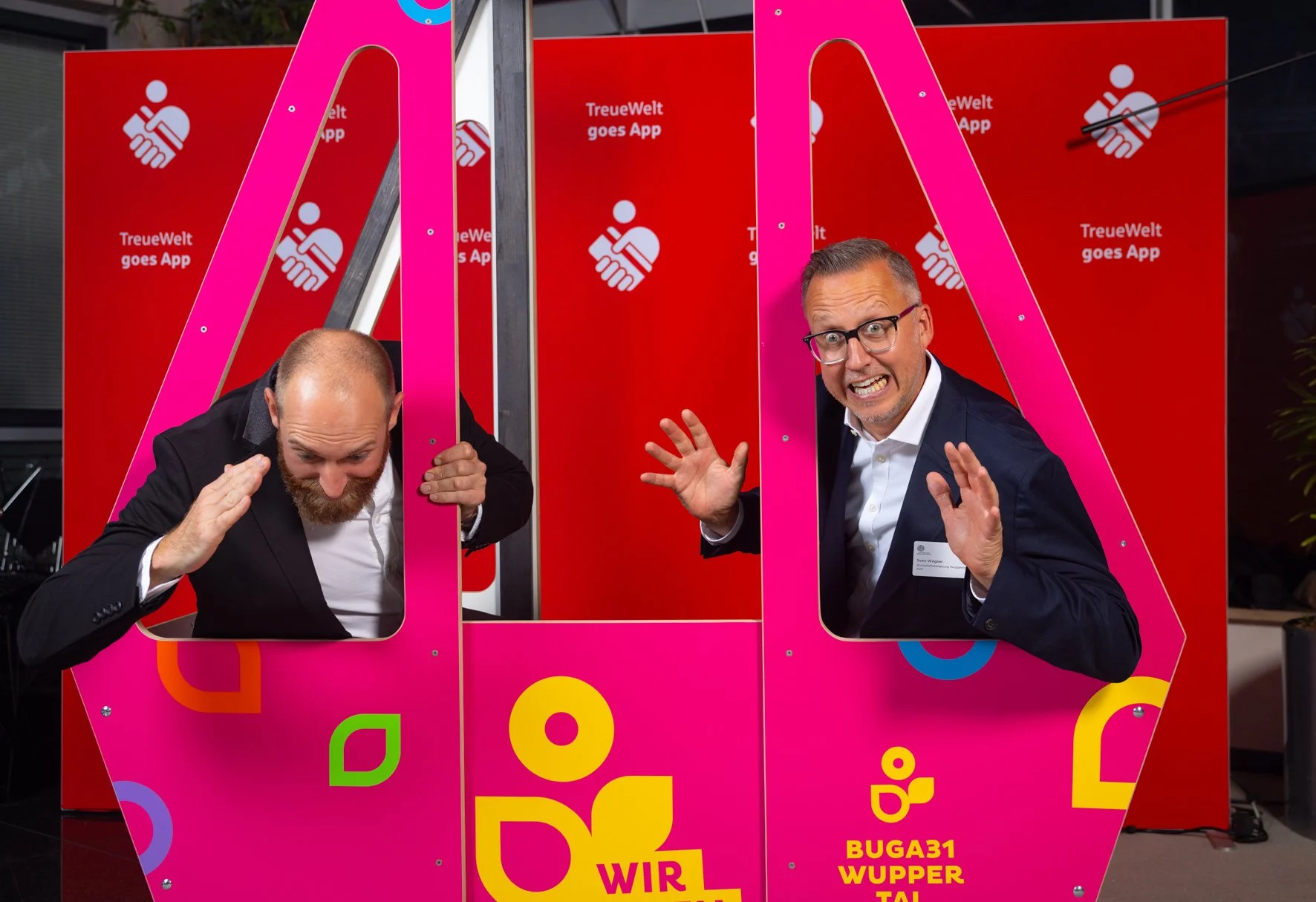 Zwei Männer, die durch eine pinkfarbene Fotokabine greifen. Der Hintergrund zeigt eine rote Wand mit dem Text "TreueWelt goes App". Der Mann auf der linken Seite hat einen Bart und trägt ein schwarzes Jackett, während der Mann auf der rechten Seite B