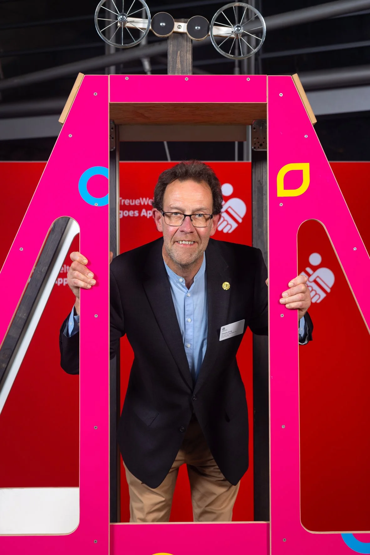 Ein Mann in formeller Kleidung steht in einem pinkfarbenen Rahmen mit einem Spielzeugauto auf dem Kopf, das wie ein Roboter aussieht, und hält die Rahmen an den Seiten.