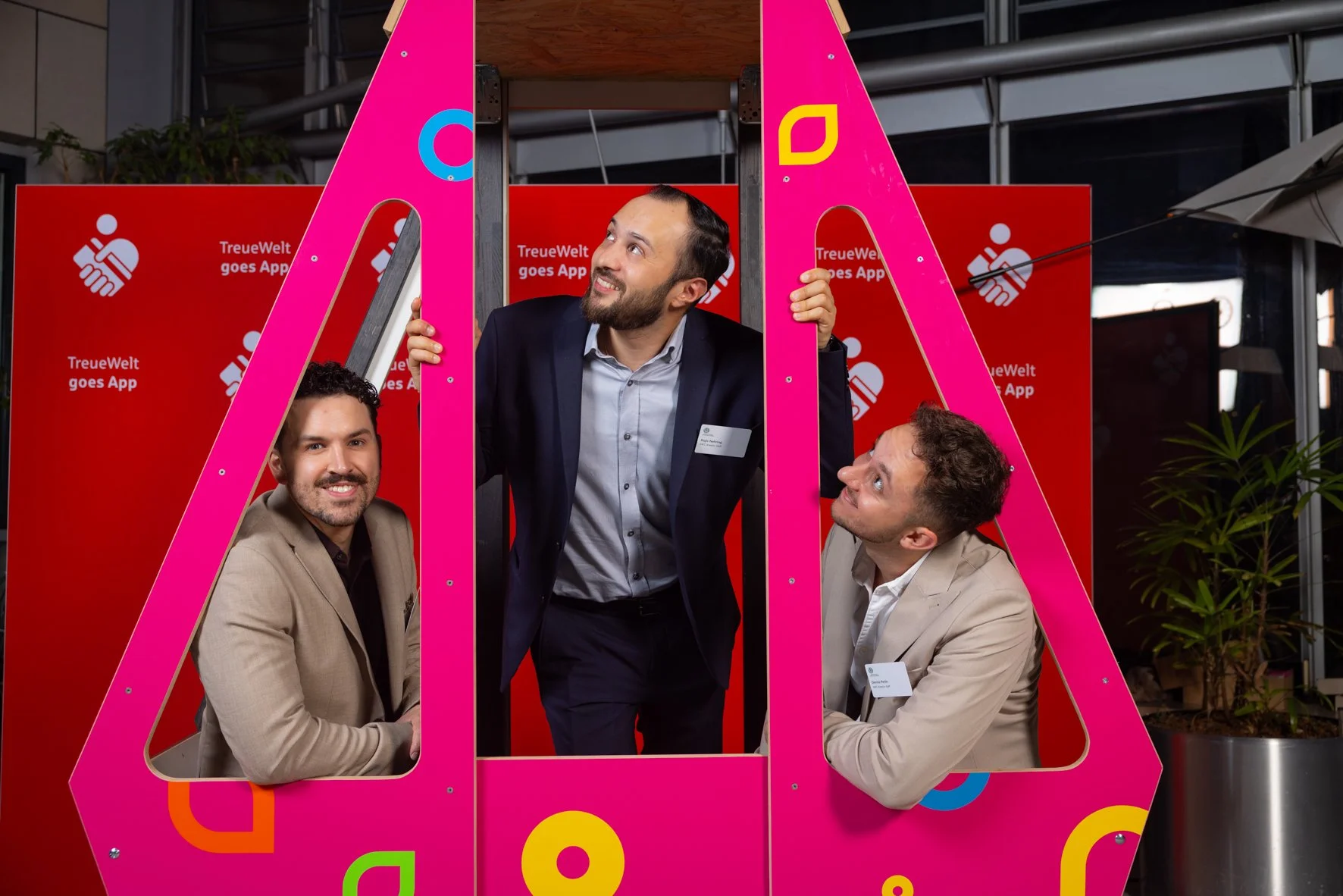 Drei Männer posieren in einer pinkfarbenen Fotokabine mit rote Hintergrund, auf dem steht 'TreueWelt goes App'.