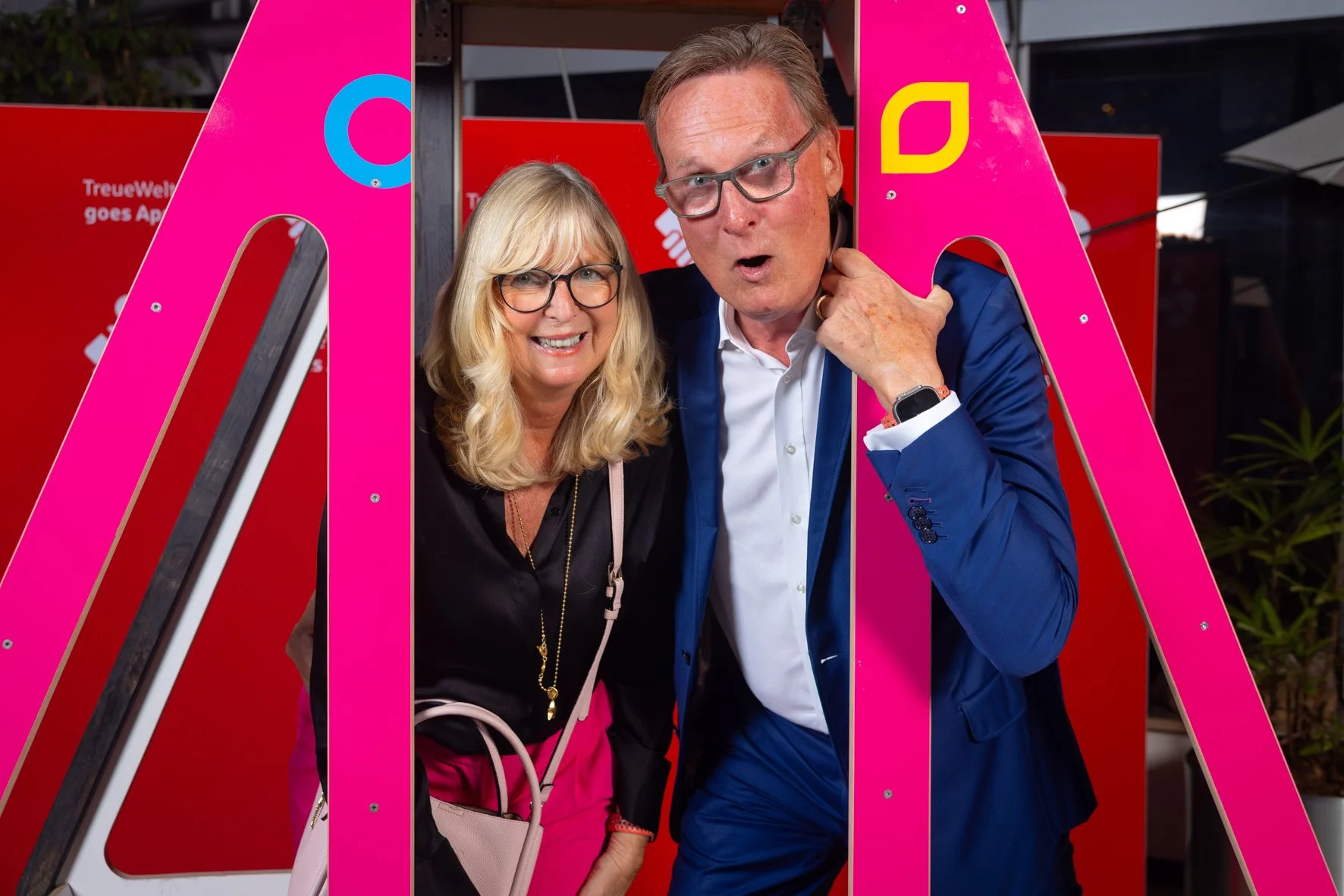 Ein Mann und eine Frau posieren hinter einem großen pinken Buchstaben auf einer roten Wand. Beide tragen Brillen und lächeln freudig.