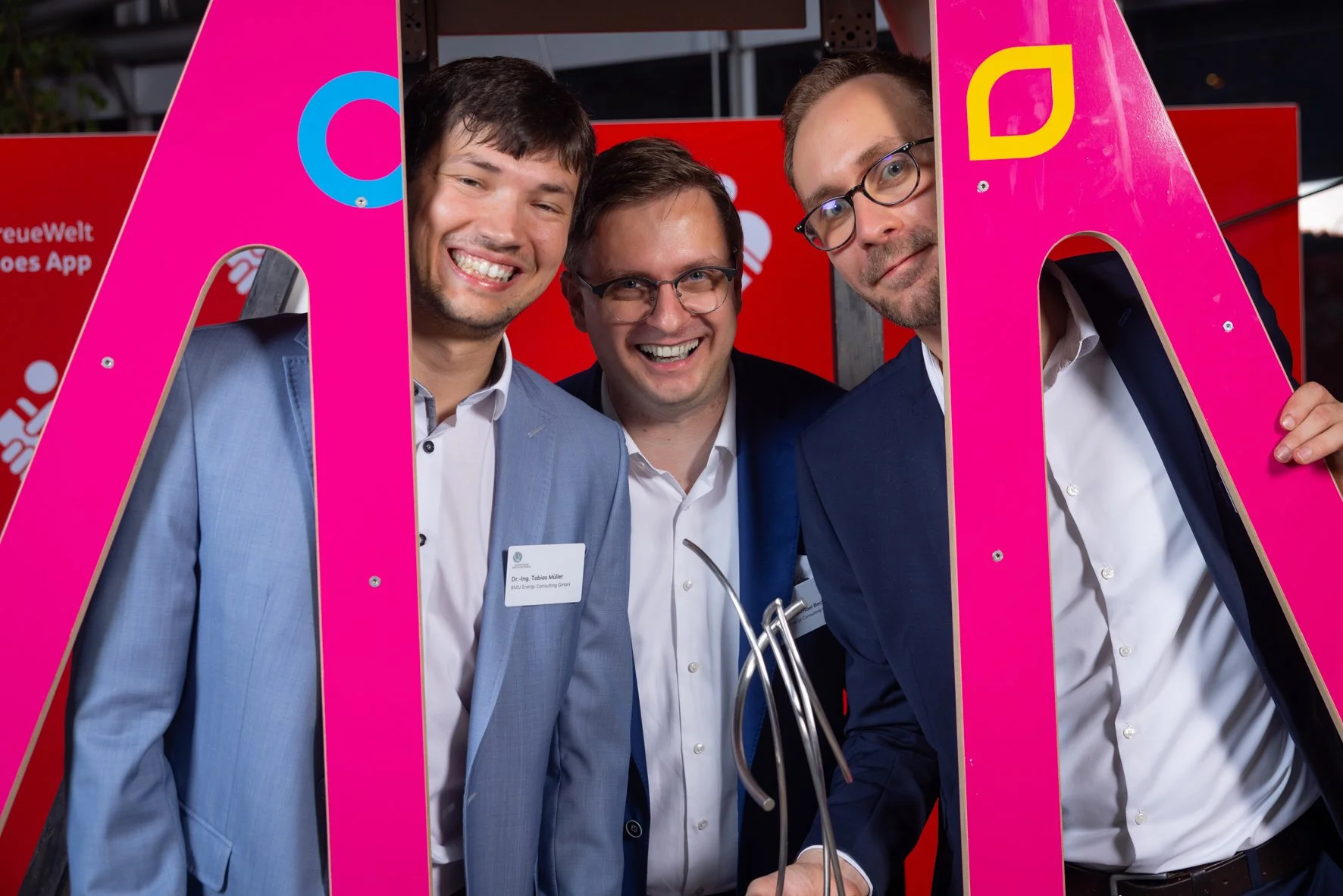 Drei Männer in Anzügen posieren hinter großen pinken Buchstaben bei einer Veranstaltung, lächeln in die Kamera.