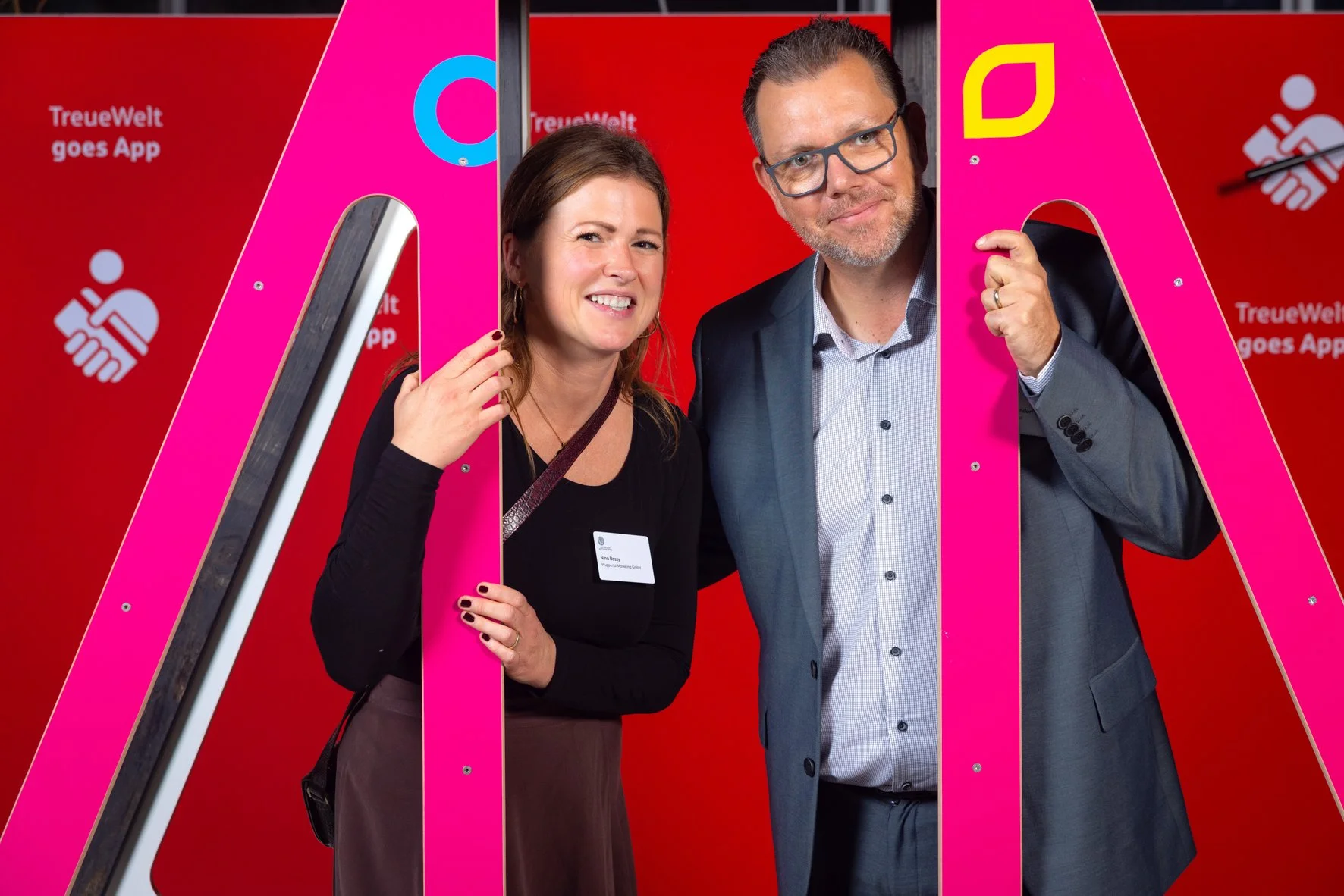 Zwei Menschen, eine Frau und ein Mann, posieren zusammen beim Foto. Sie stehen hinter einem großen rosa Buchstaben 'M' und haben Lächeln auf den Gesichtern vor einem roten Hintergrund. Die Frau trägt eine schwarze Bluse mit Namensschild, der Mann trä