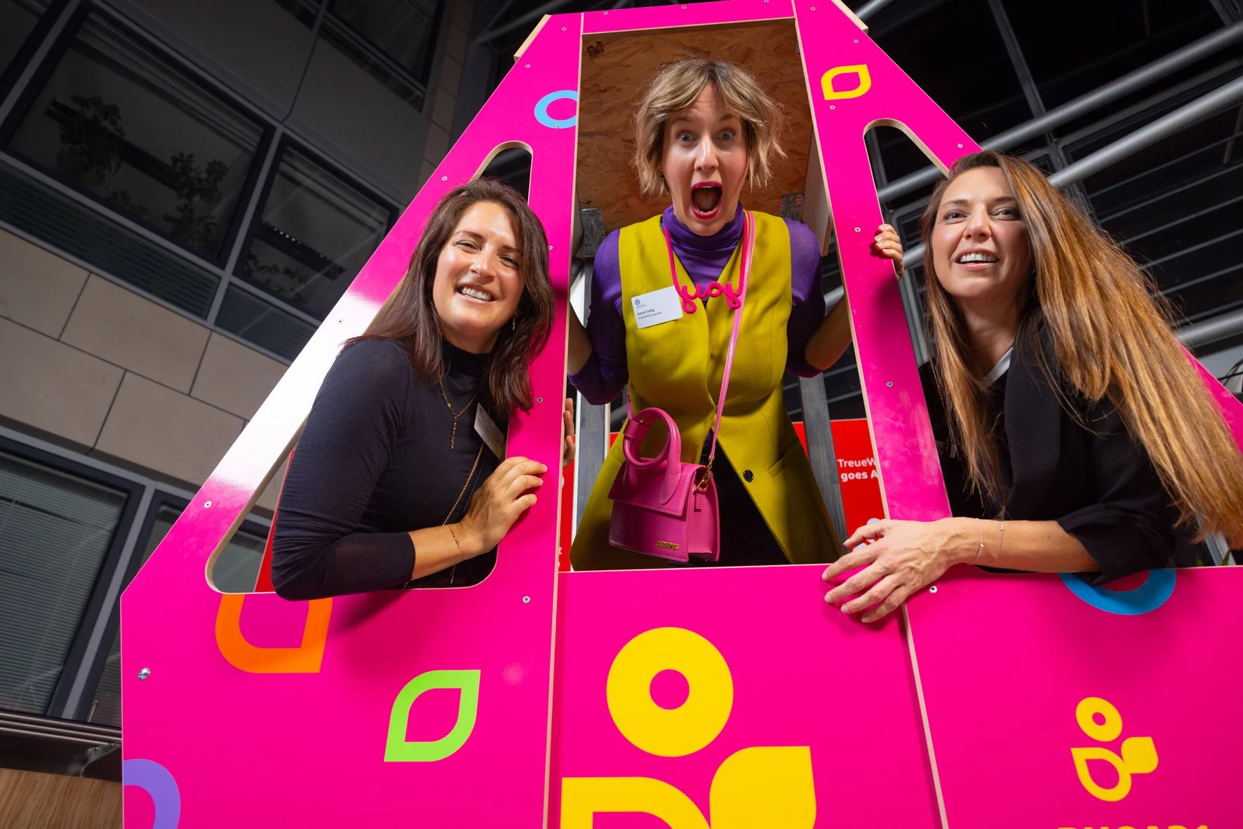 Drei Frauen posieren lachend auf einer pinkfarbenen und bunten Fotokabine im modernen Gebäude.