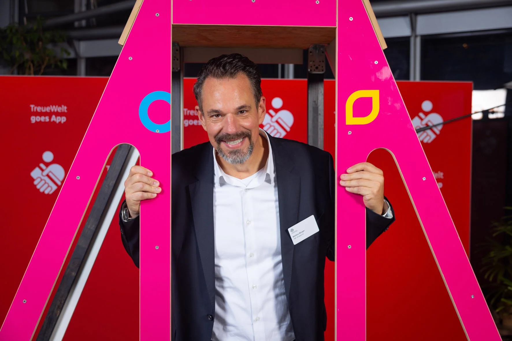 Ein Mann in einem schwarzen Anzug und weißem Hemd steht hinter einer pinkfarbenen, offenen Buchstaben-Installation. Er lächelt und hält die Rahmen der Buchstaben. Im Hintergrund sind rote Plakate mit weißen Logos und Texten sichtbar.