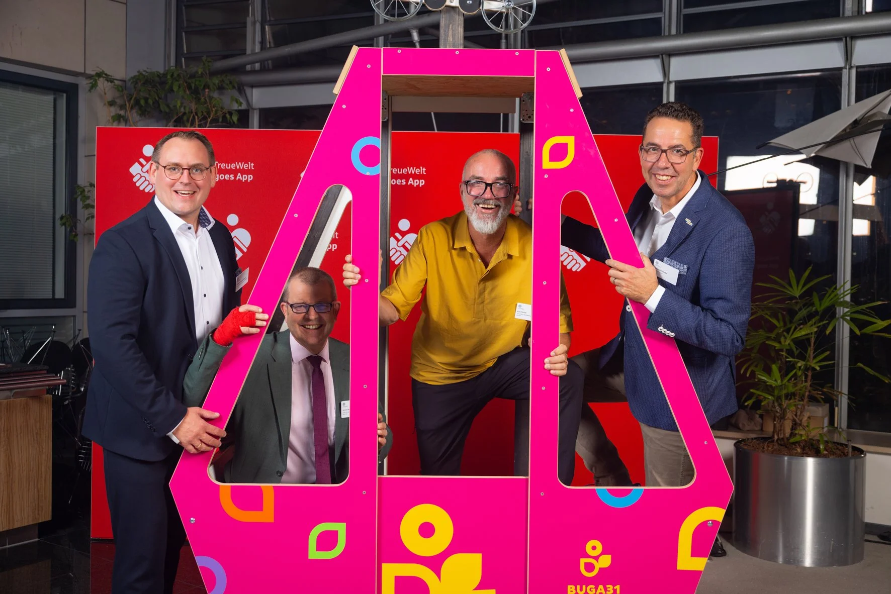 Vier Männer posieren vor einer roten Wand mit dem Text 'FreueWelt goes App'. Sie stehen hinter einem großen pinken Rahmen in Form eines Smartphones. Die Männer lachen und smile. Es scheint eine professionelle Veranstaltung oder ein Fotoshooting zu se