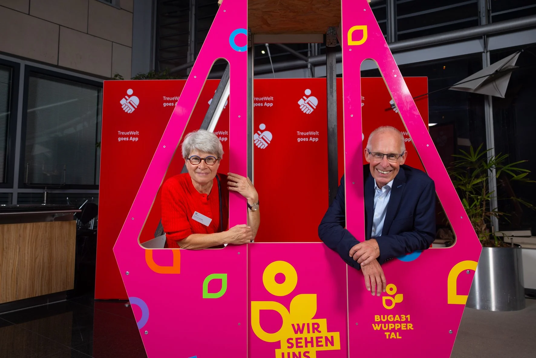 Zwei Personen, eine ältere Frau und ein älterer Mann, lachen und posieren hinter einer pinkfarbenen Kunstinstallation mit bunten grafischen Elementen. Im Hintergrund sind rote Werbetafeln mit weißem Text und Symbolen.