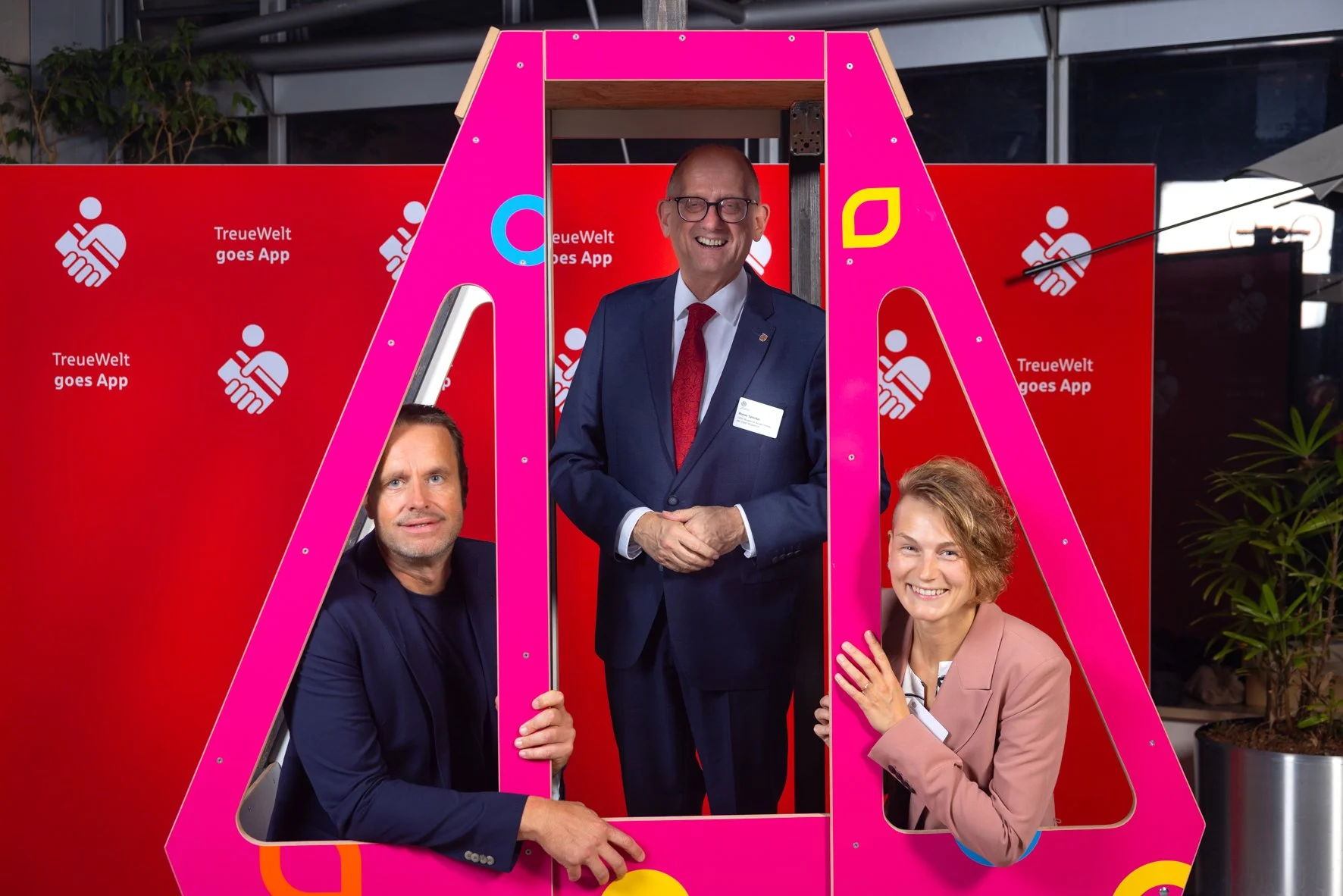 Drei Menschen posieren vor einem roten Hintergrund mit der Aufschrift 'TreueWelt goes App'. Sie stehen in einer pinkfarbenen Rahmenkonstruktion, die an ein Papierhandtuch erinnert. Der zentrale Mann trägt einen dunkelblauen Anzug mit roter Krawatte u