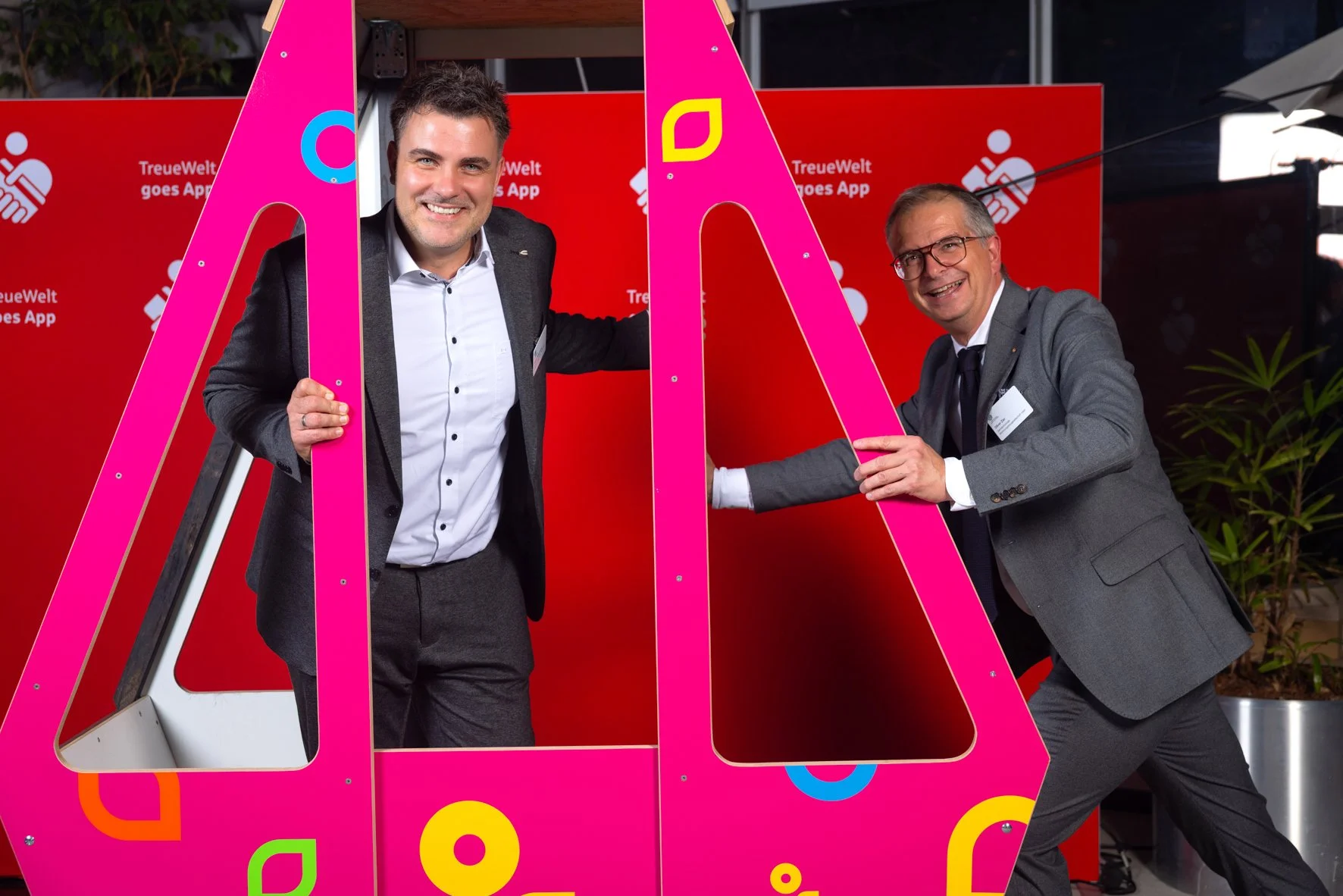 Zwei Männer in formeller Kleidung spielen bei einer Veranstaltung mit einem großen pinkfarbenen Rahmen, vor einem roten Hintergrund mit dem Text 'TreueWelt goes App' und einem Icon. Beide lachen und sehen glücklich aus.
