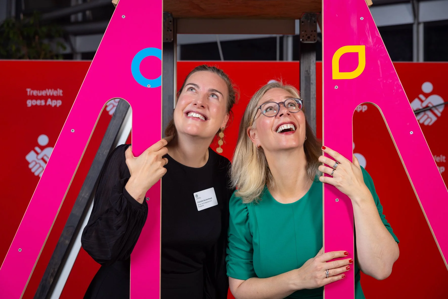 Zwei Frauen lächeln und schauen nach oben, während sie durch eine pinke Rahmenöffnung posieren, bei einer Veranstaltung mit einem roten Hintergrund. Eine Frau trägt eine schwarze Jacke und eine blaue Namensschild, die andere trägt ein grünes Kleid un