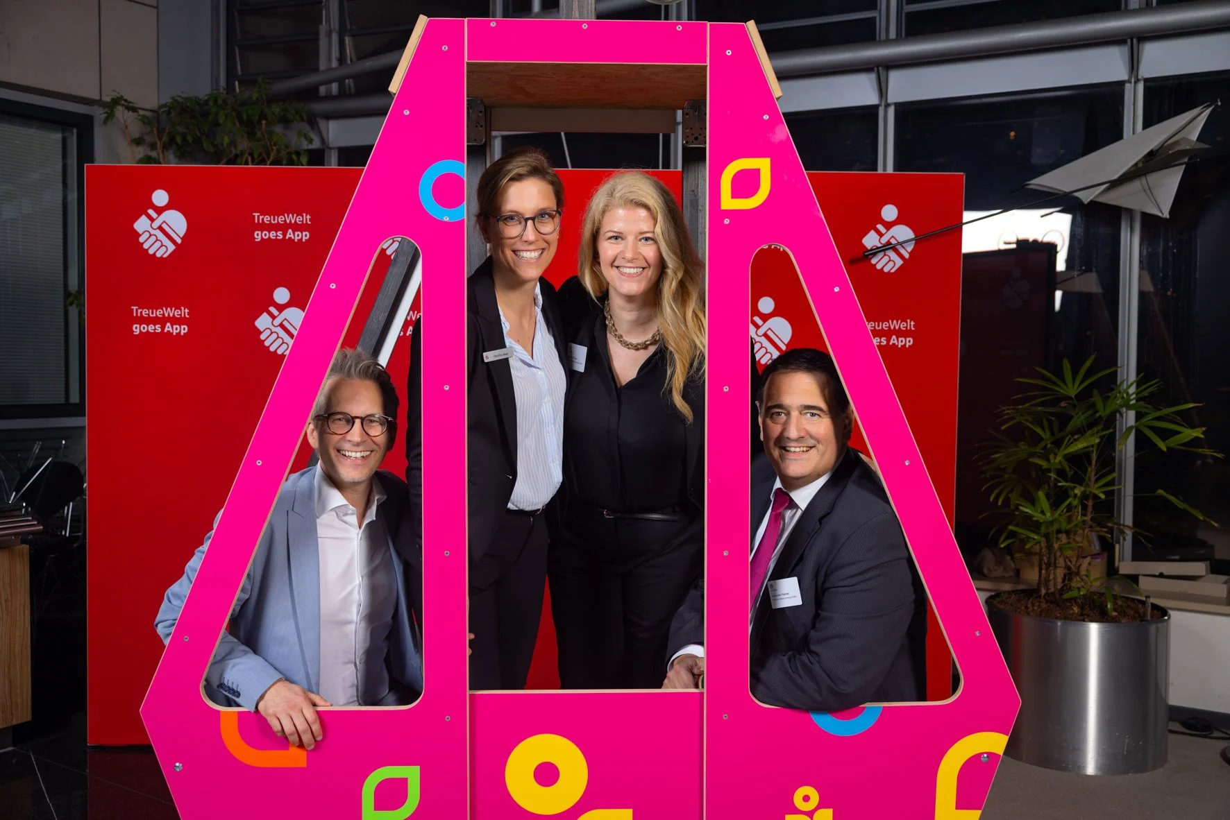 Vier Menschen bei einer Veranstaltung, die in einem pinkfarbenen Fotoautomat stehen, in der Nähe einer roten Wand mit dem Logo und der Aufschrift 'TreueWelt goes App'.