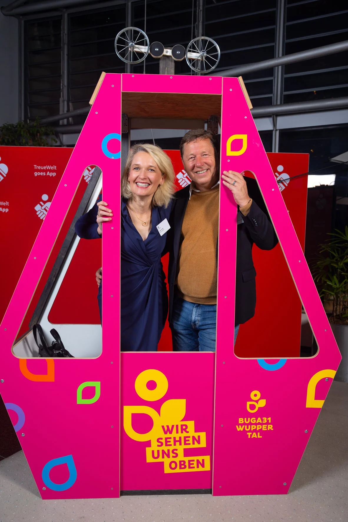 Zwei Menschen posieren bei einer Fotokabine, die in rosa mit bunten Mustern gestaltet ist. Sie lachen und wirken fröhlich.