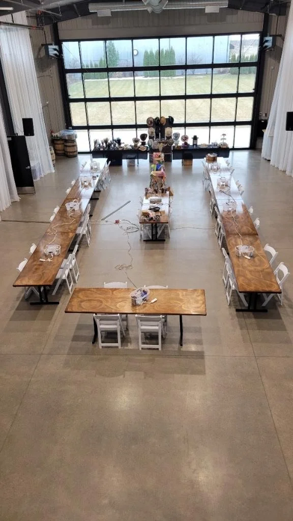 Long rectangular tables arranged in a U-shape inside a large, modern event space with a concrete floor and large windows at the back. The tables are covered with white tablecloths and set with plates, glasses, and cutlery, suggesting preparations for
