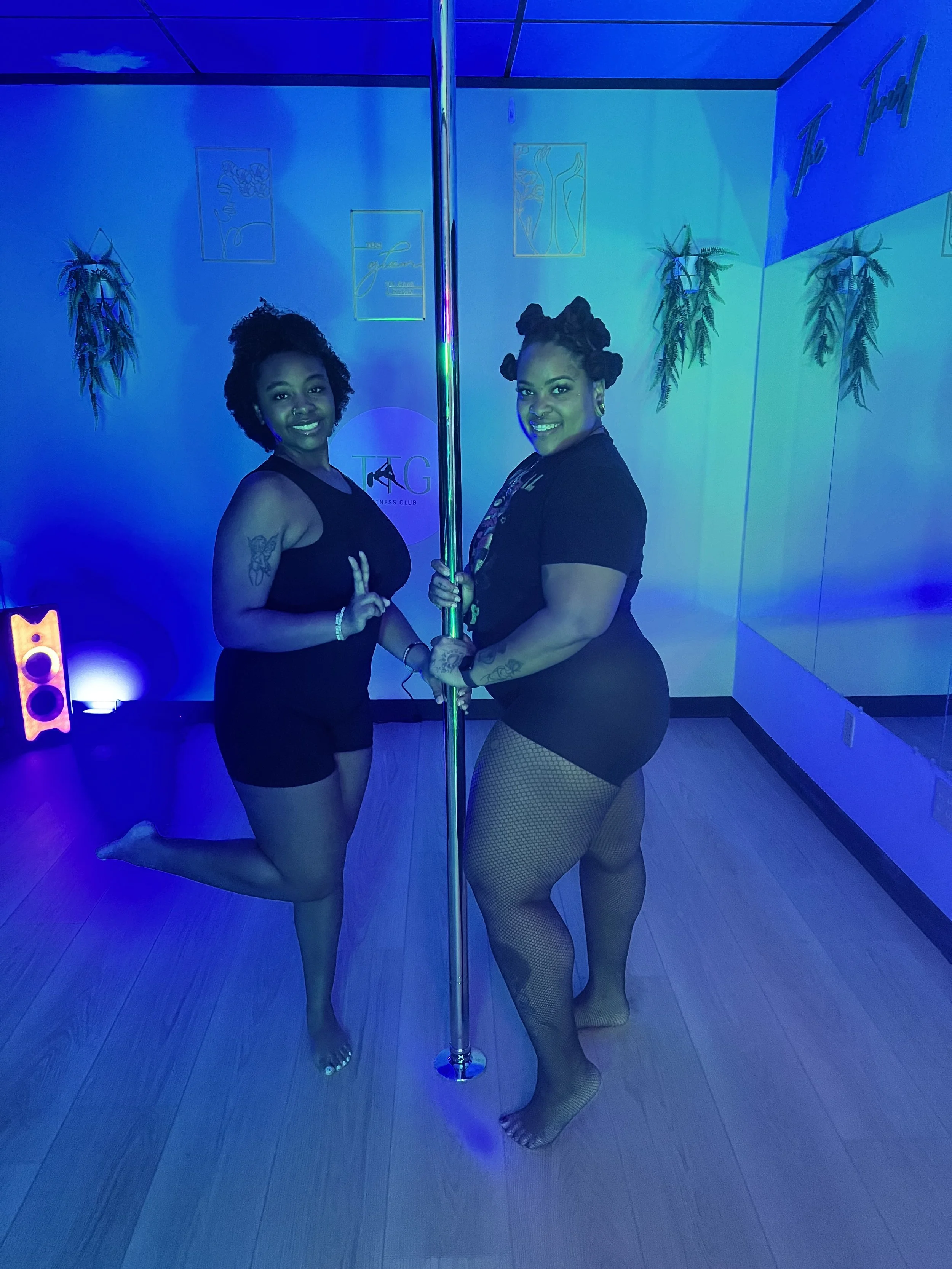 Two women posed on a dance pole in a dimly lit dance studio with blue lighting. They are smiling and holding the pole, with wall decorations in the background.