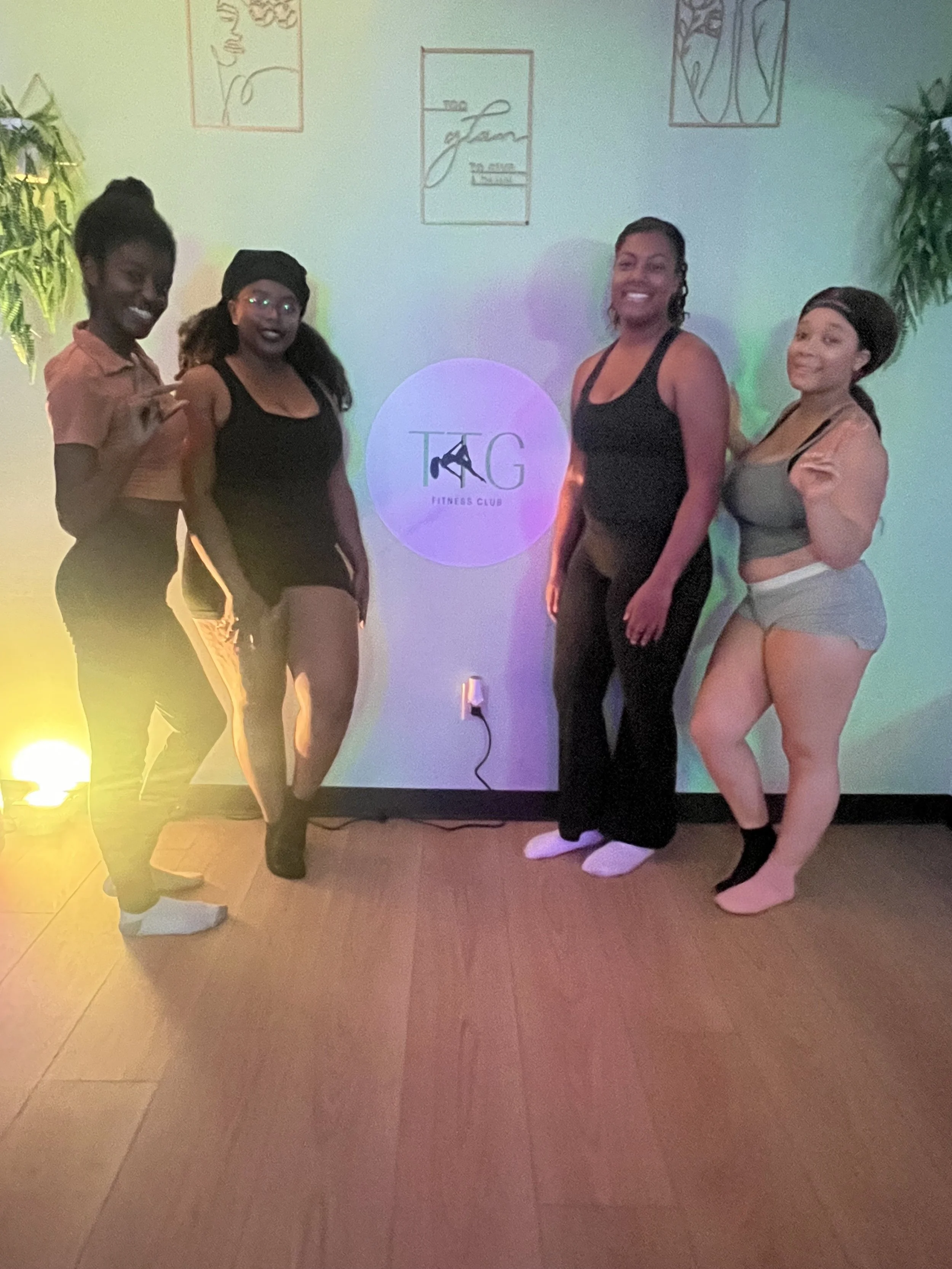 Four women standing in front of a wall with a logo that says 'TAG Fitness Club,' with colorful lights and plants in the background.