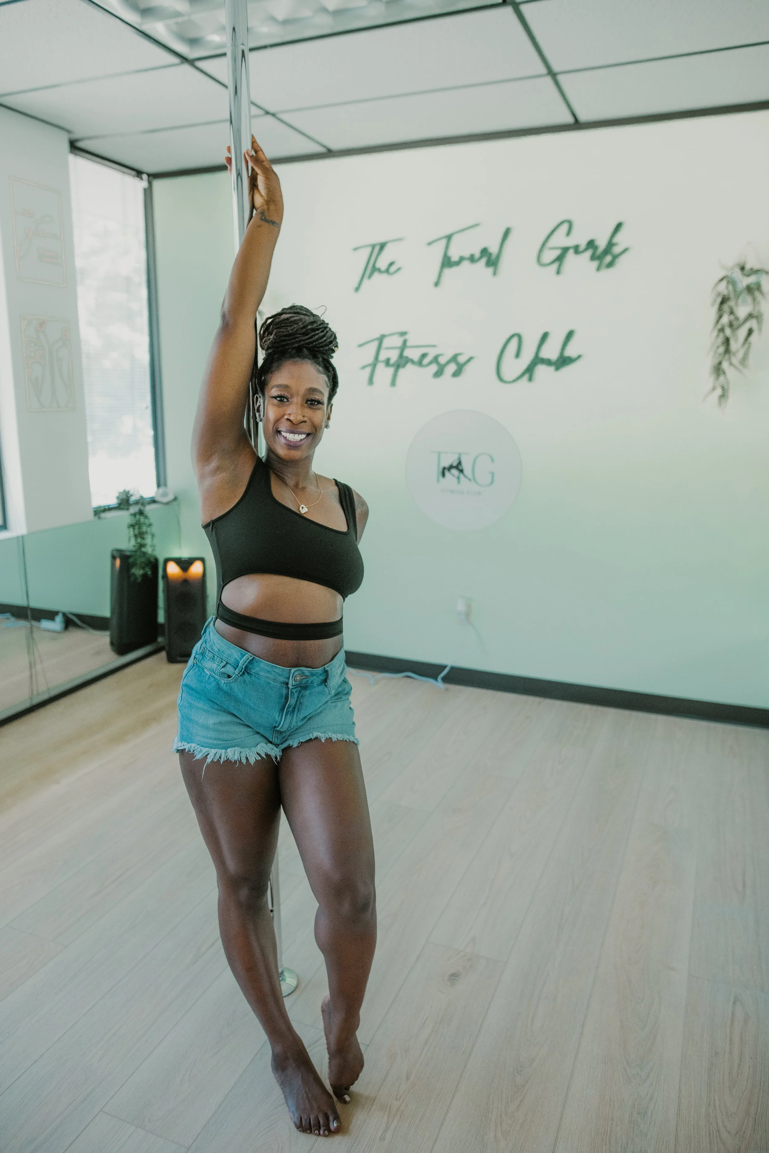 A woman practicing pole dance indoors, smiling while holding onto a pole. She is wearing a black crop top and denim shorts, with a fitness studio in the background that has a mirror, a logo on the wall, and some plants.