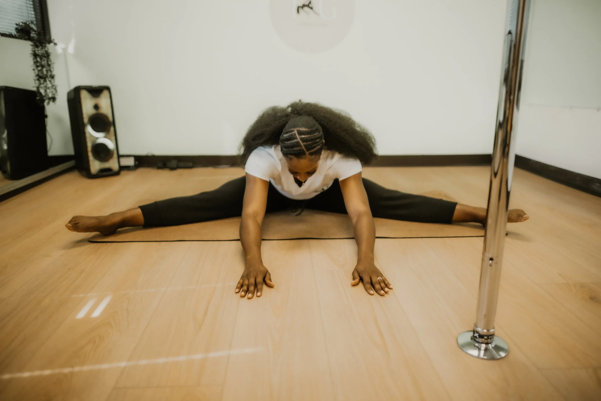 A woman practicing split yoga pose on a wooden floor, stretching with her hands on the ground and a ballet barre nearby.