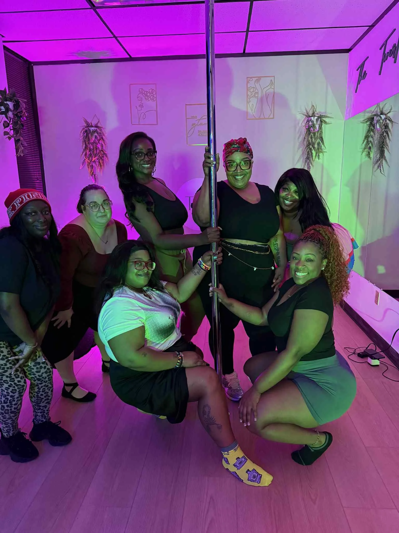Group of women at a pole dancing studio, some kneeling, some standing, with purple lighting and wall art in the background.