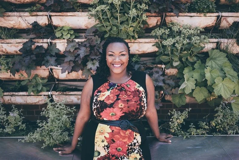 Woman in a floral dress smiling in front of a vertical garden wall with various plants.