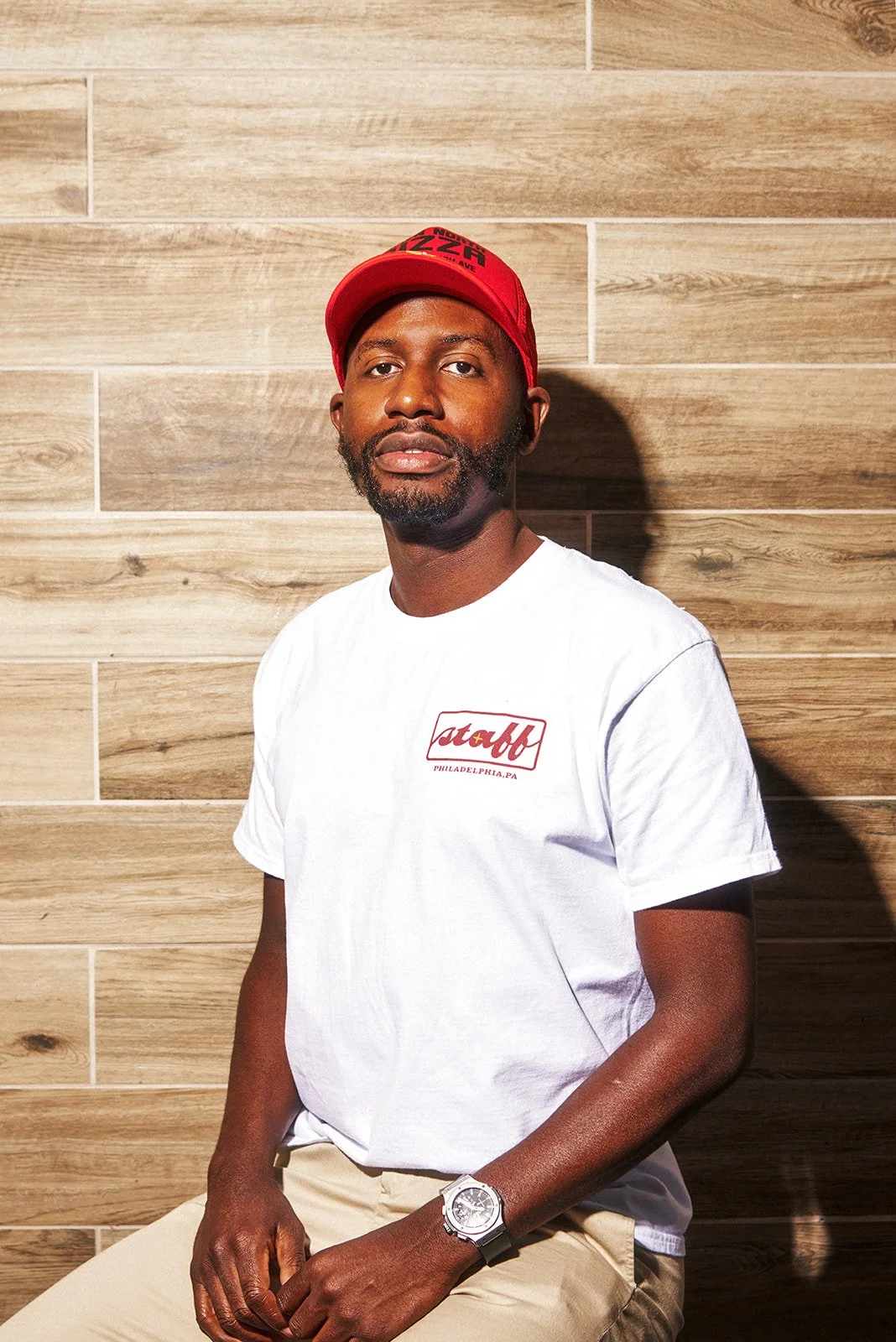Man in white T-shirt and red cap sitting against a wooden wall.