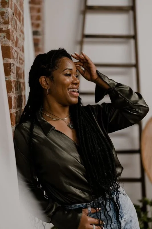 Smiling woman with long hair leaning against a brick wall, wearing a dark blouse and jeans, with a blurred ladder in the background.