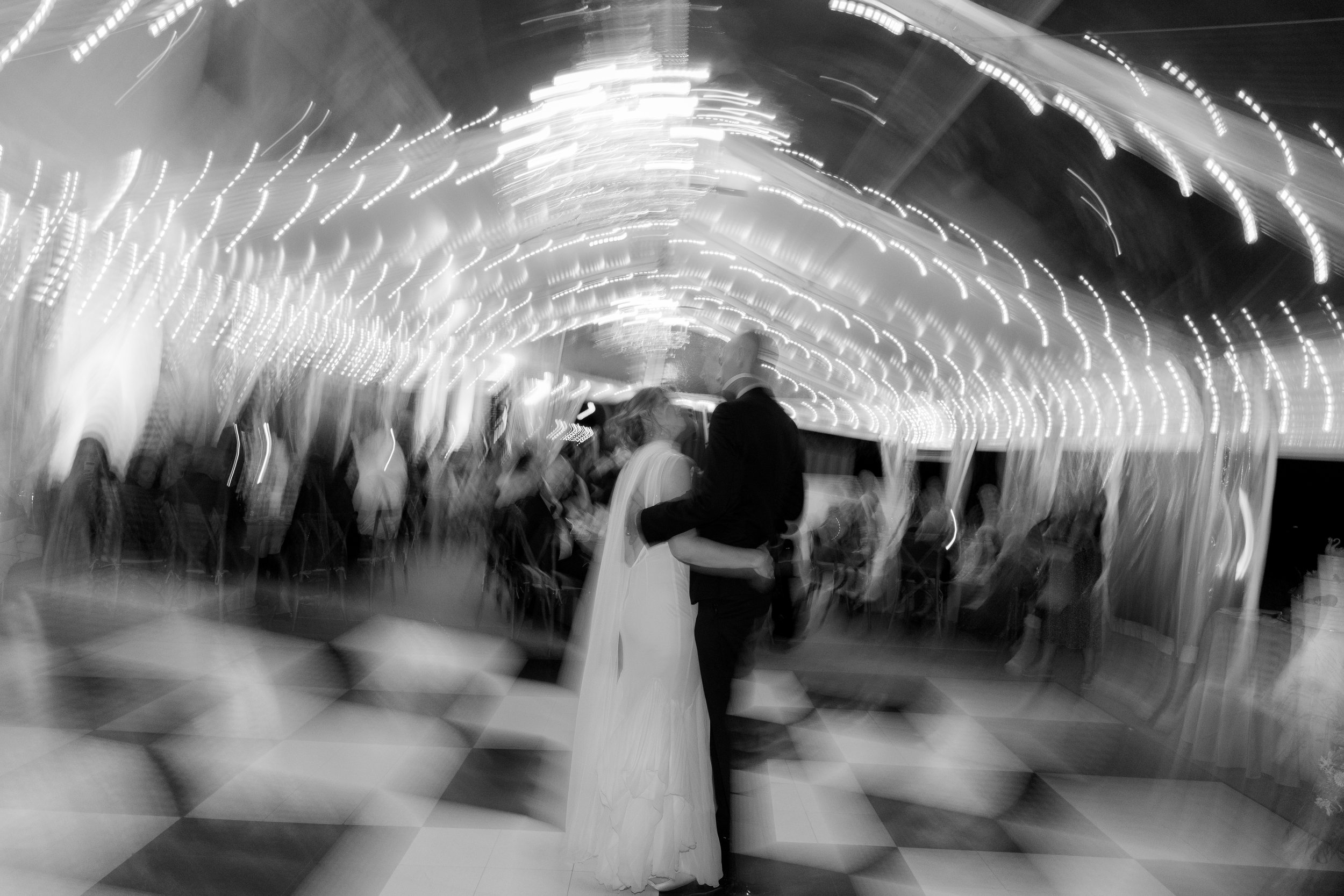 A bride and groom share a dance at their wedding reception with guests seated around them under string lights, captured in black and white with motion blur.