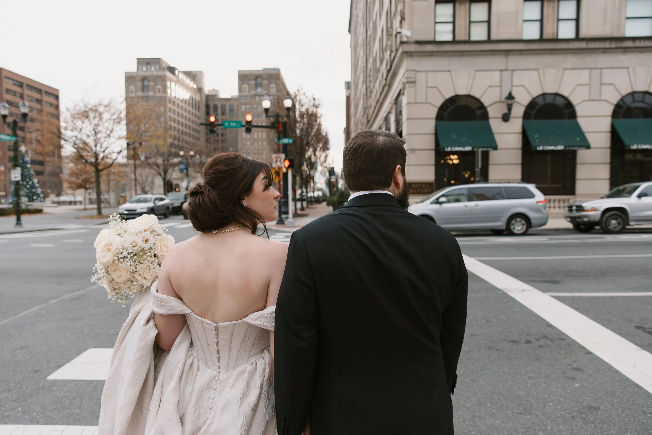 A newly married couple stands at a city street intersection, with the woman in a wedding dress holding a bouquet of white roses and a man in a tuxedo.