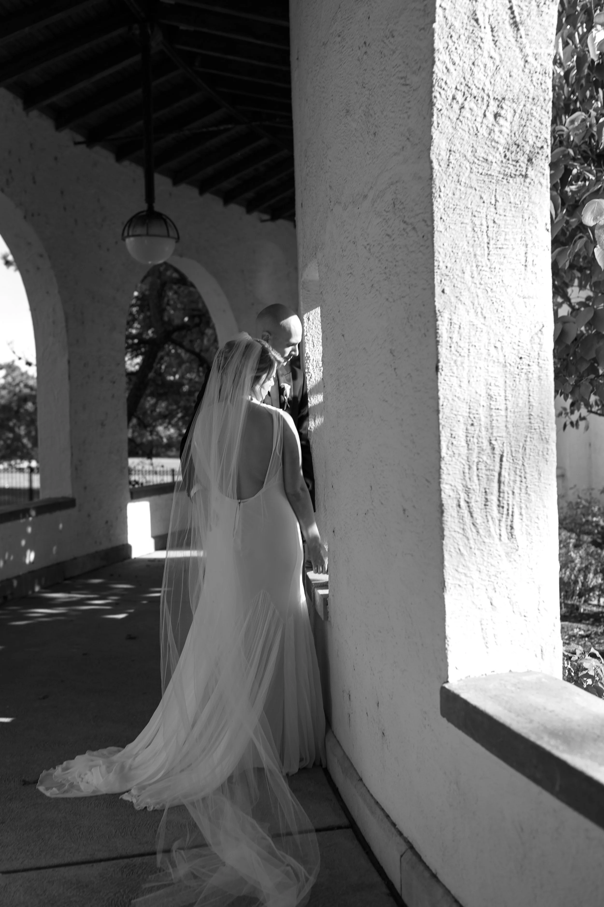 Black and white photo of a bride and groom with their heads bowed together, standing under an arched walkway with sunlight casting shadows.