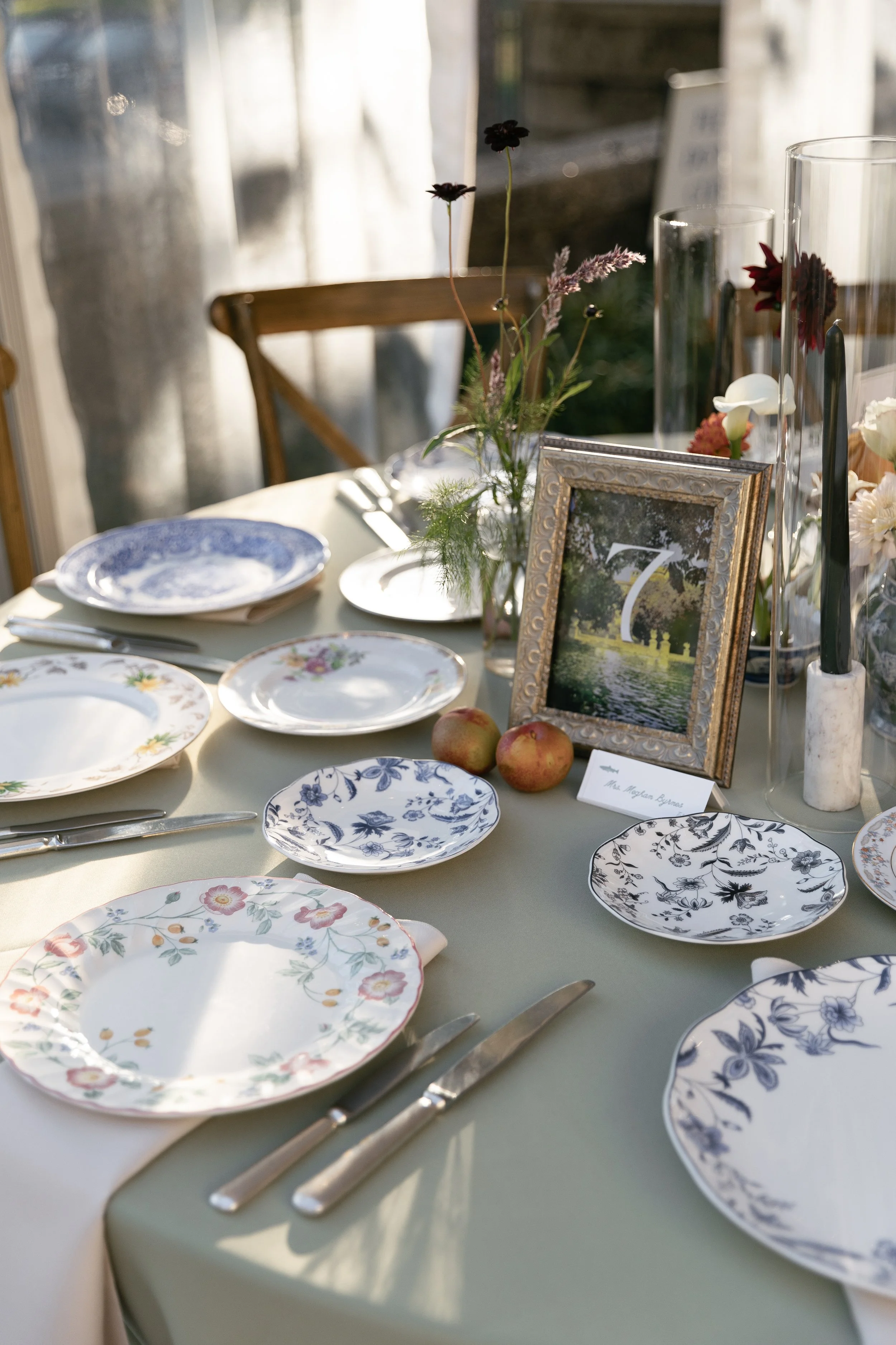 A table set for a meal with floral-patterned plates, silverware, and a centerpiece with flowers, apples, and picture frames, with sunlight illuminating the scene.