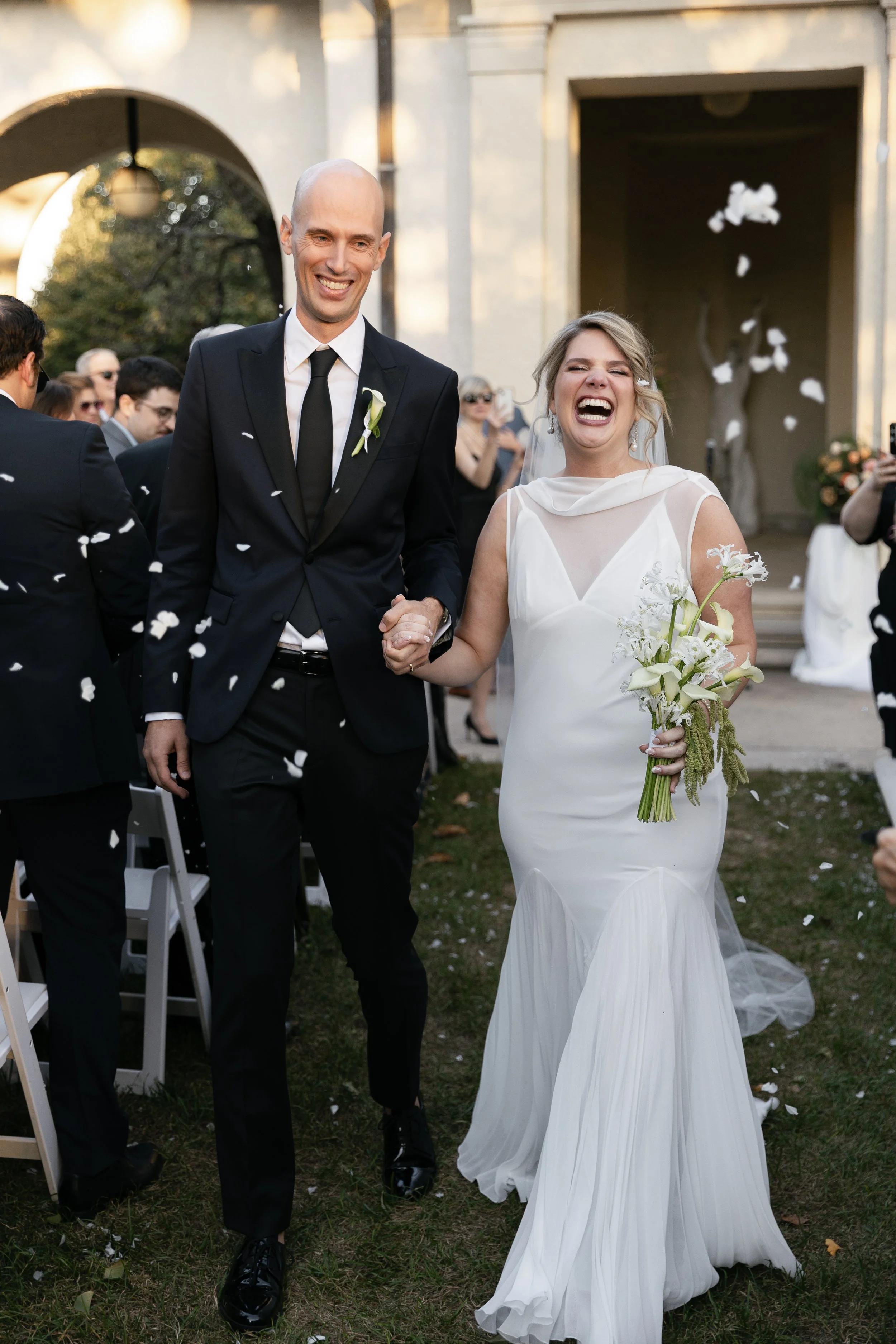 A bride and groom walking together, holding hands, while celebrating their wedding outdoors. The bride is wearing a white wedding dress and holding a bouquet of white flowers, and the groom is dressed in a black suit with a white boutonniere. They ar