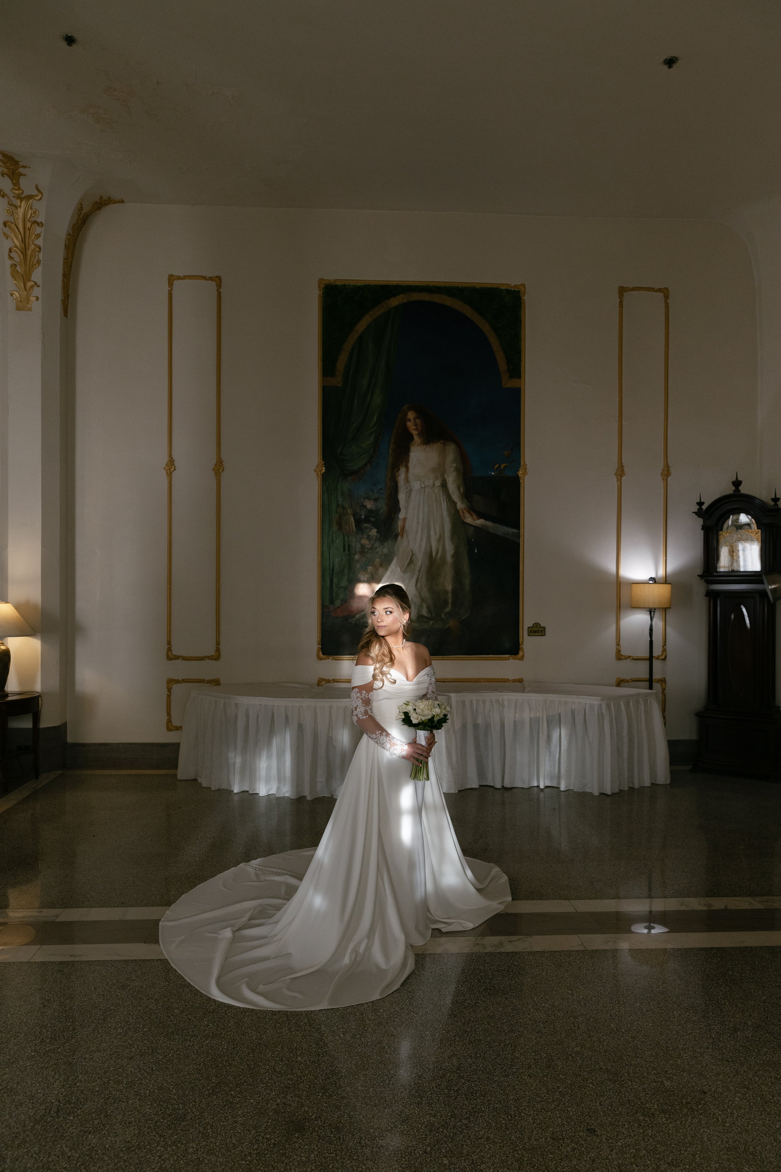 A bride in a white wedding dress holding a bouquet of white flowers standing in a gallery-like setting with large painting behind her and warm lighting.