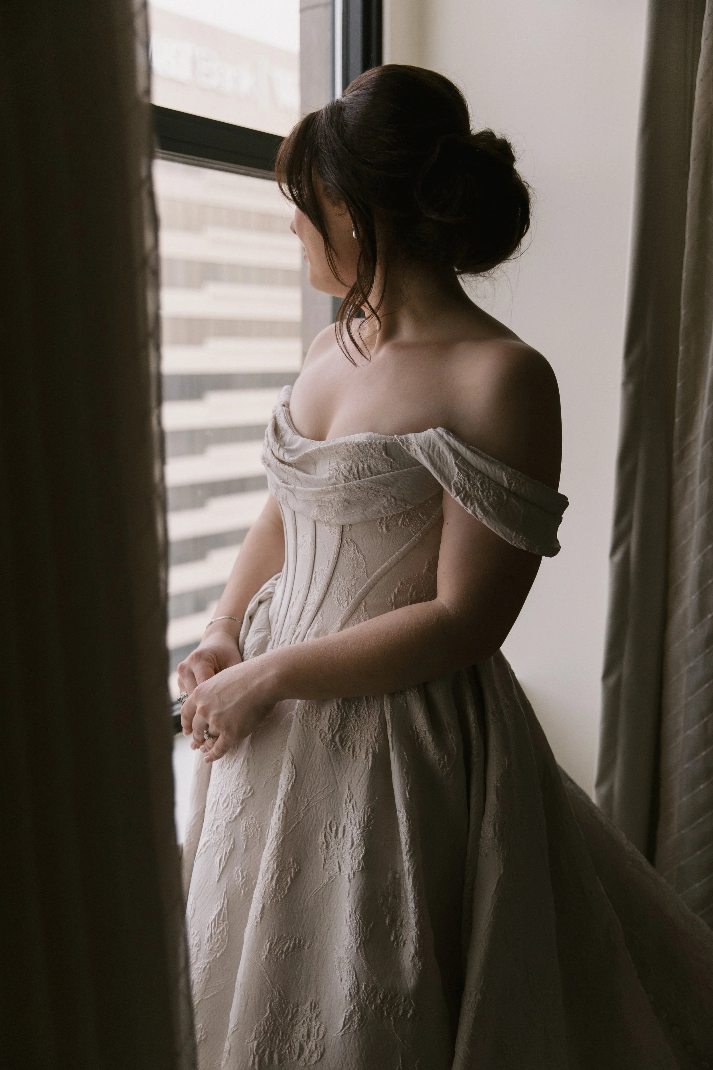 A woman in an off-shoulder, textured wedding dress stands by a window, looking away, with her hands clasped in front of her.