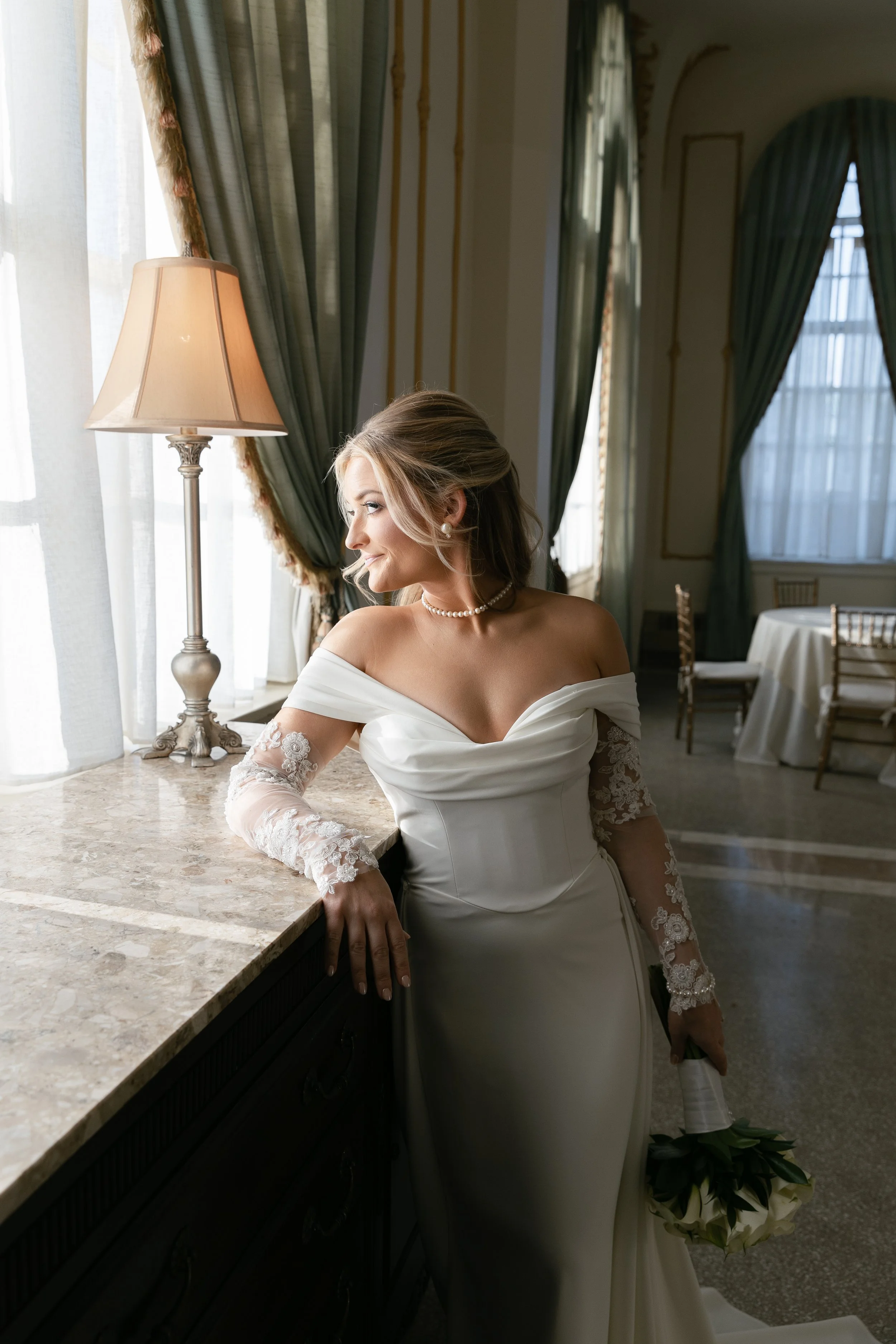 A bride in a white wedding dress with lace sleeves, holding a bouquet, standing by a window with green and white curtains, in an elegant room with high ceilings and large windows.