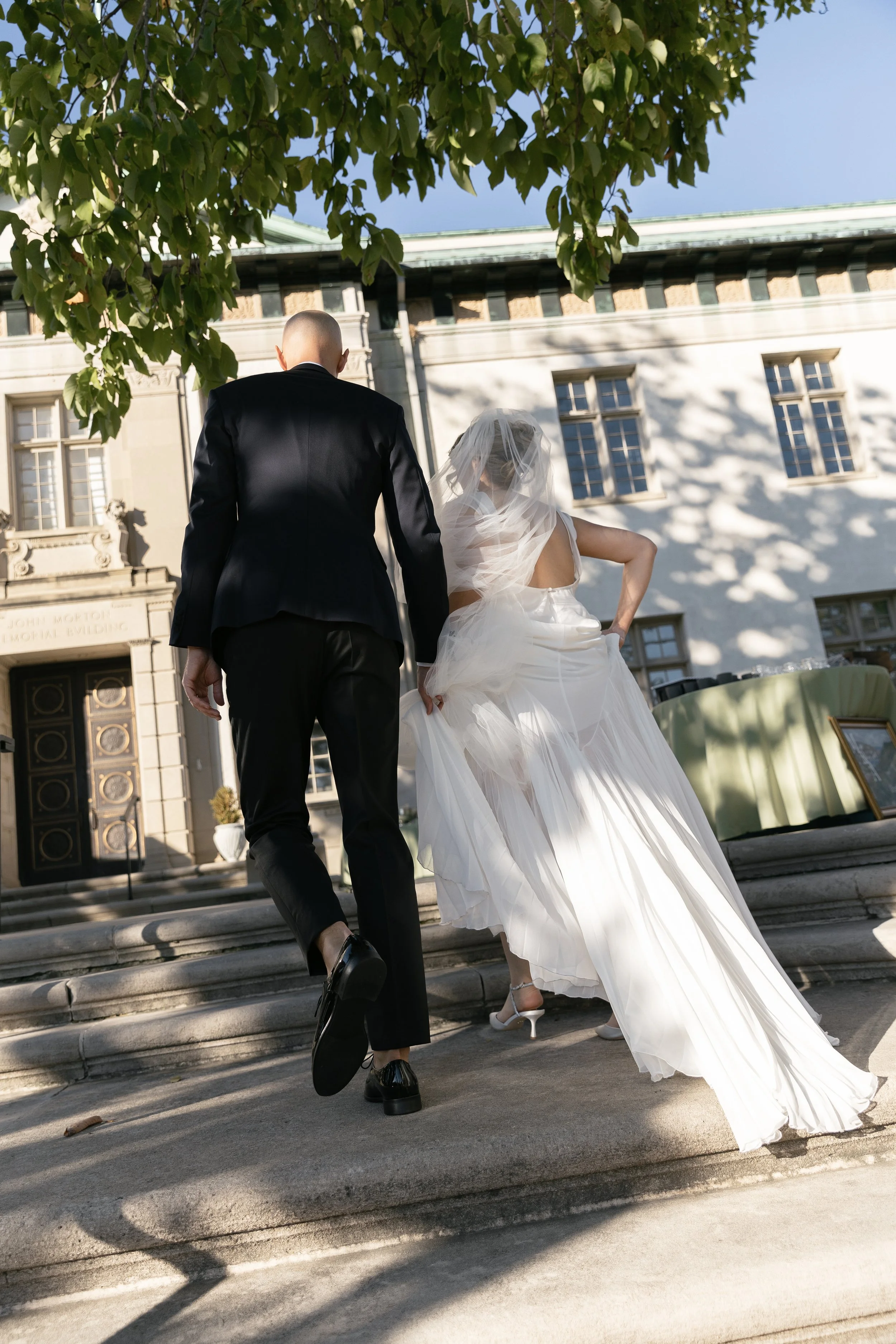 Bride and groom walking up steps outside of a building, with the groom in a black suit and the bride in a white wedding dress with veil.