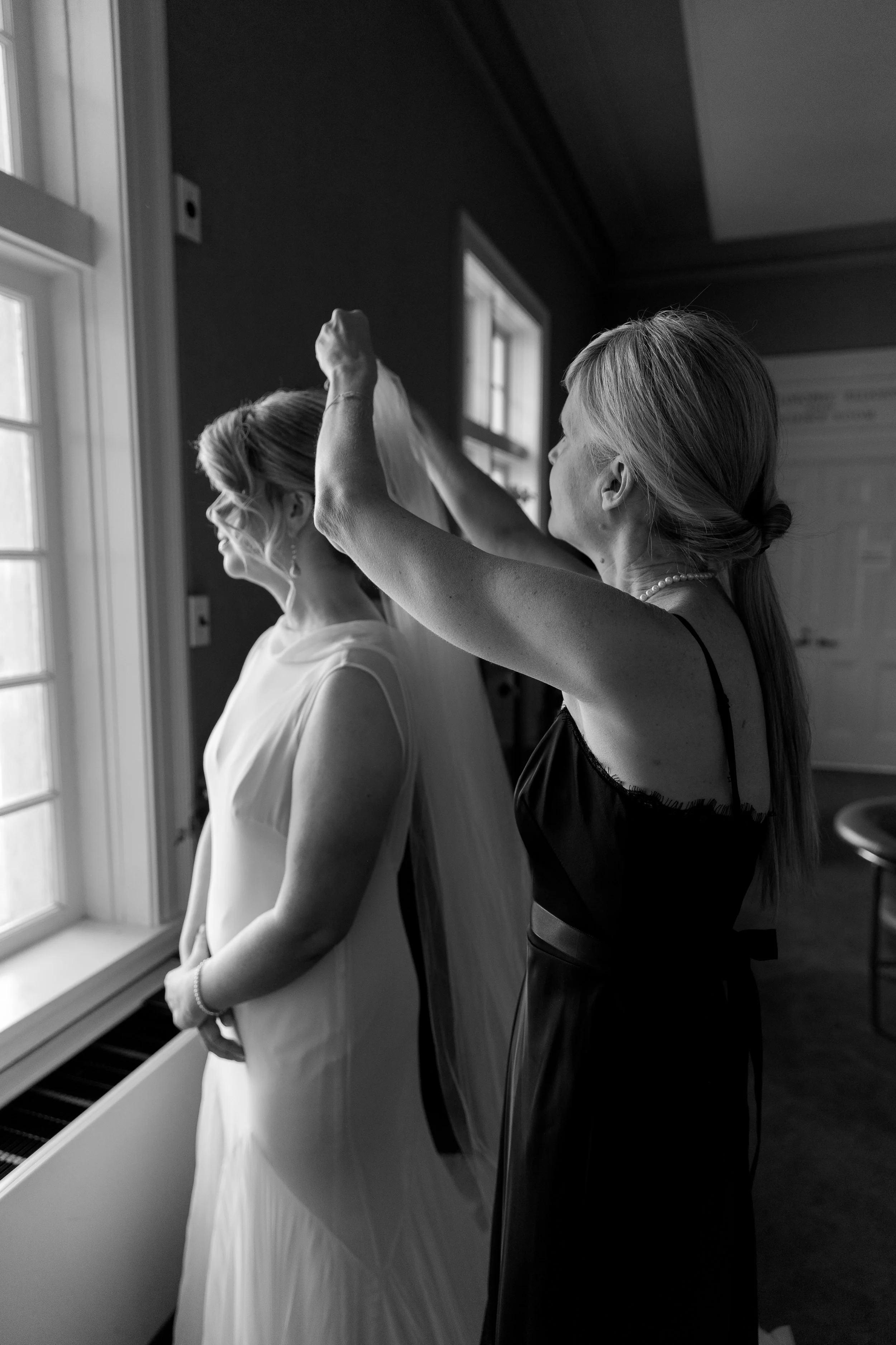 A woman helping another woman put on a wedding veil in a room with windows.