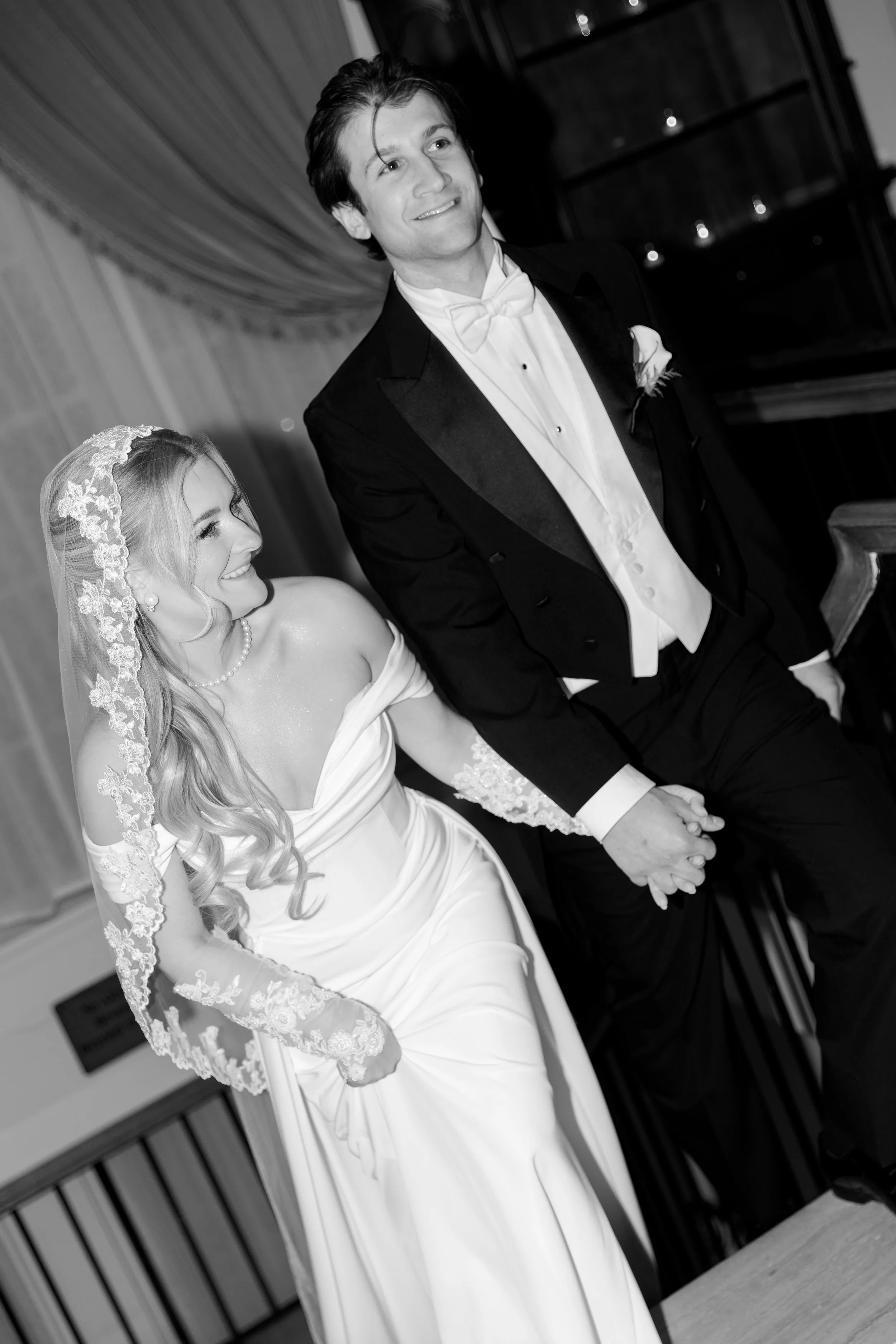 A black and white photo of a bride and groom holding hands, sitting on a piano bench during their wedding ceremony.