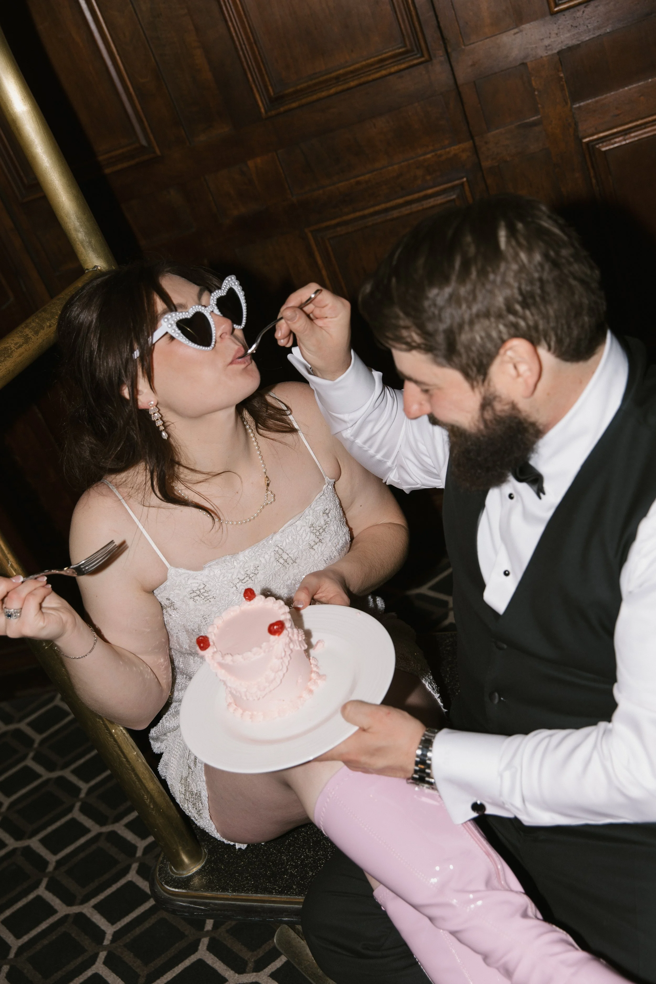 Woman in white lace dress with heart-shaped sunglasses being fed cake by a man in a tuxedo at a celebration, holding a pink cake with red cherries.
