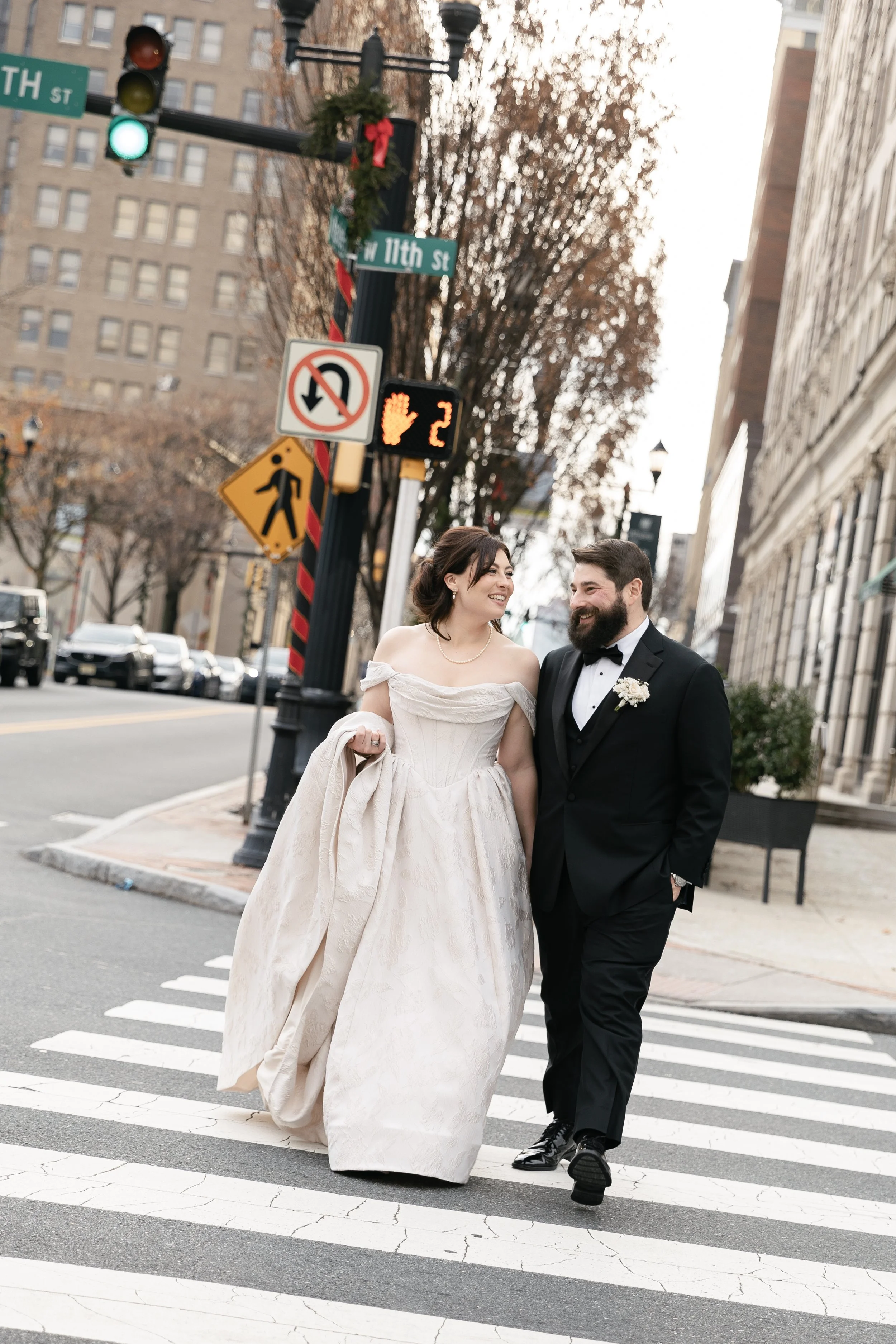 A newlywed couple in wedding attire walking across a crosswalk in an urban setting. The bride is in a white off-the-shoulder wedding gown holding part of her dress, and the groom is in a black tuxedo with a boutonniere, both smiling at each other.