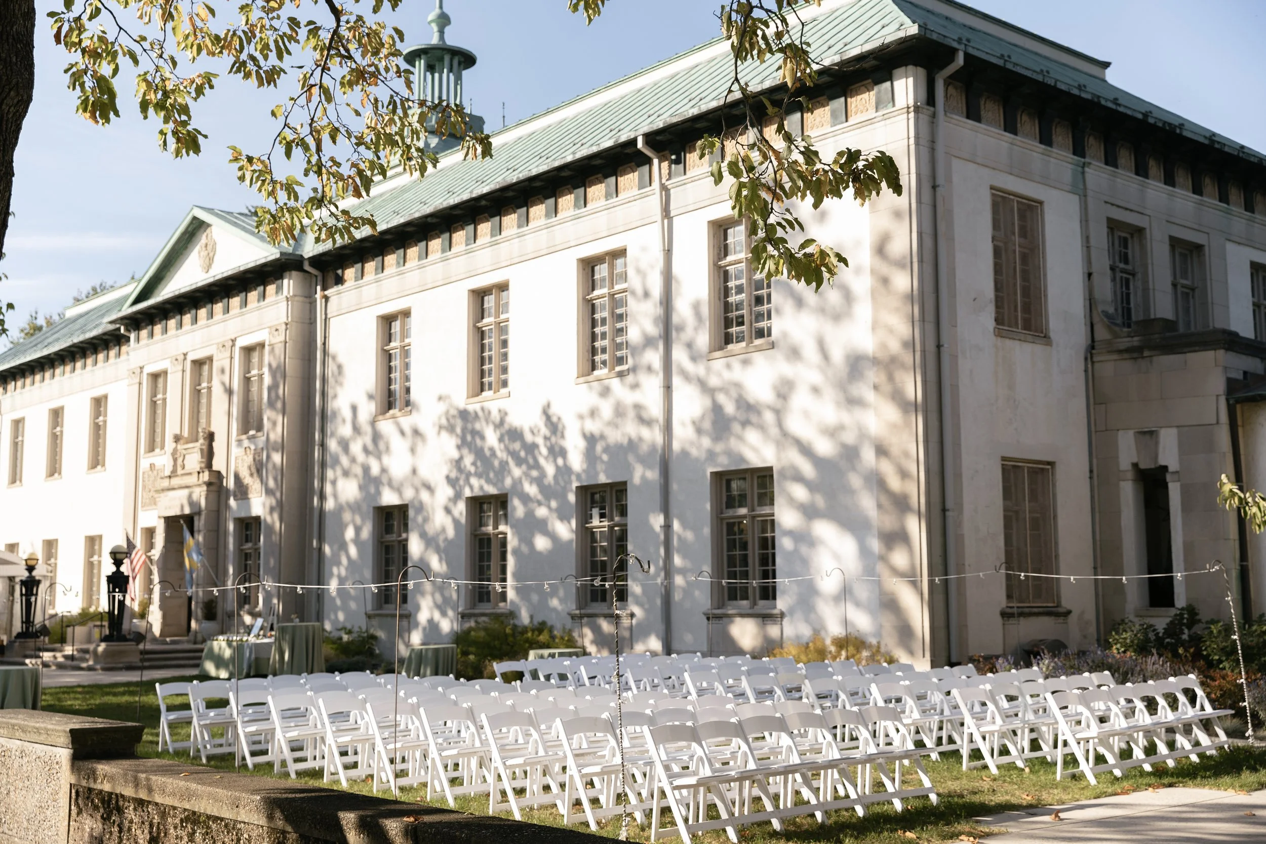 An outdoor scene in front of a large historic building with multiple windows and a green roof, scenic setup for an event with many white folding chairs arranged on the grass, and tables with green tablecloths underneath string lights, shaded by trees