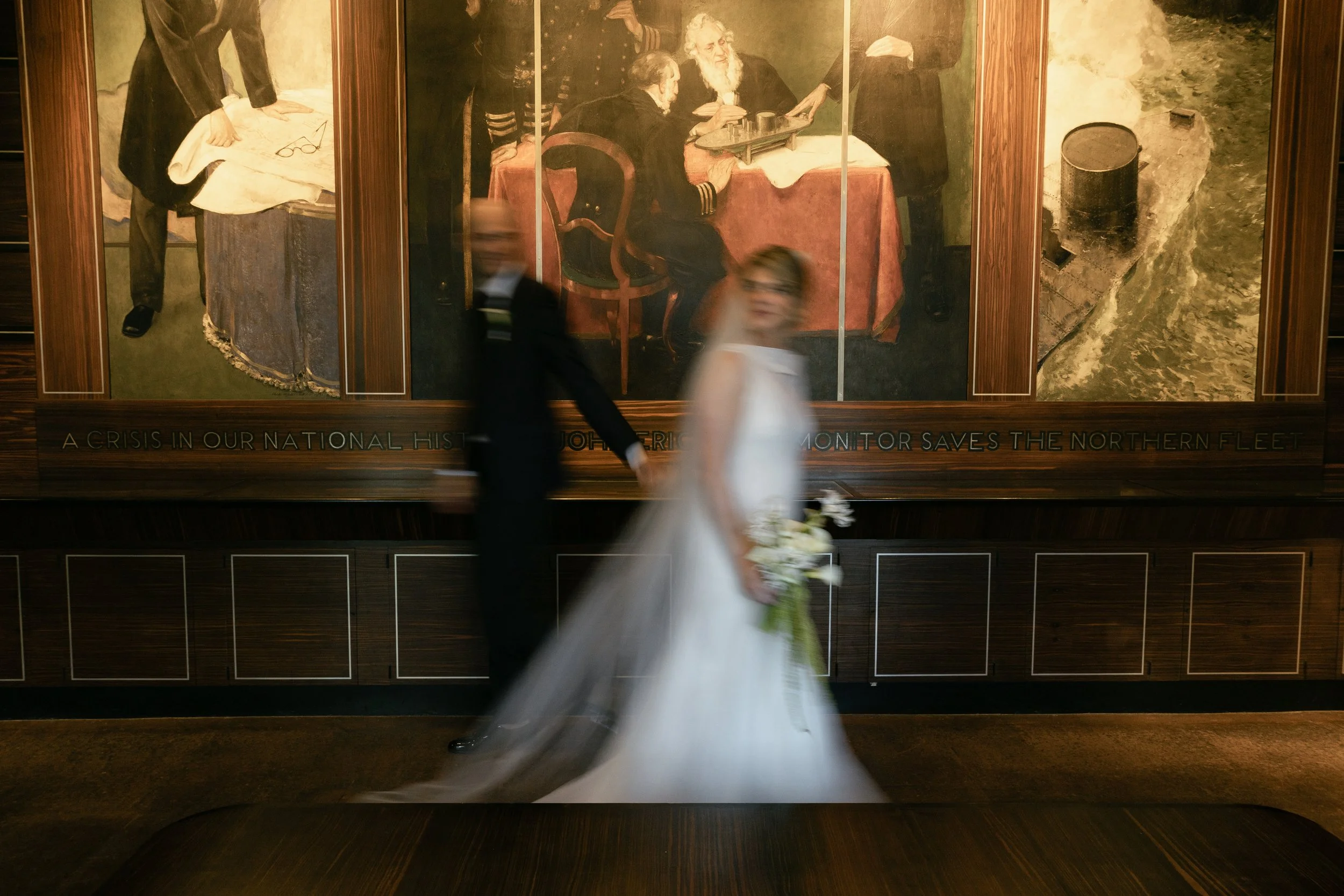 A blurry wedding couple holding hands, with the bride in a white dress holding a bouquet, walking past a large historical painting in a museum or gallery.