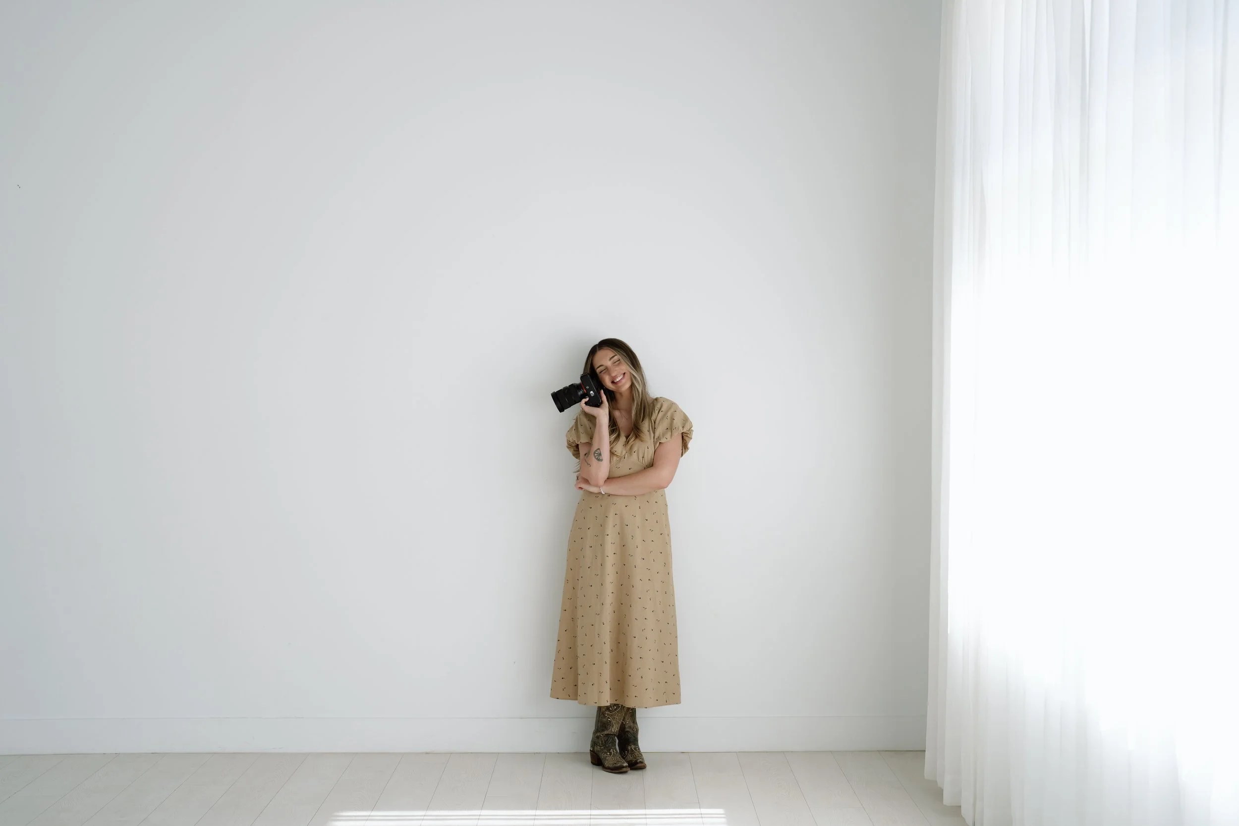 girl standing against white wall holding camera, smiling with eyes closed, wearing tan dress and cowboy boots