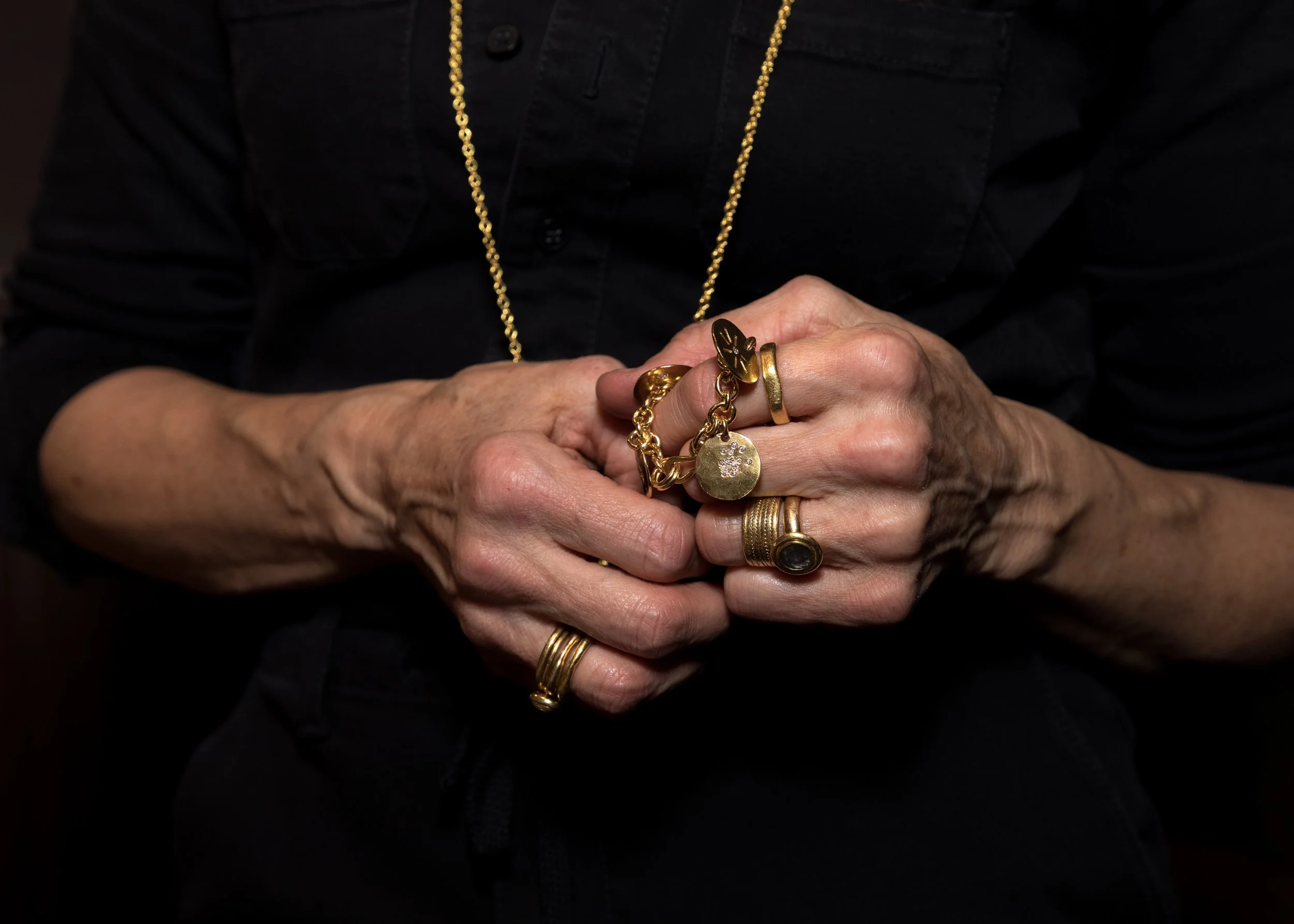 Close-up of aged hands holding jewelry, including rings and gold chains, against a black shirt background.