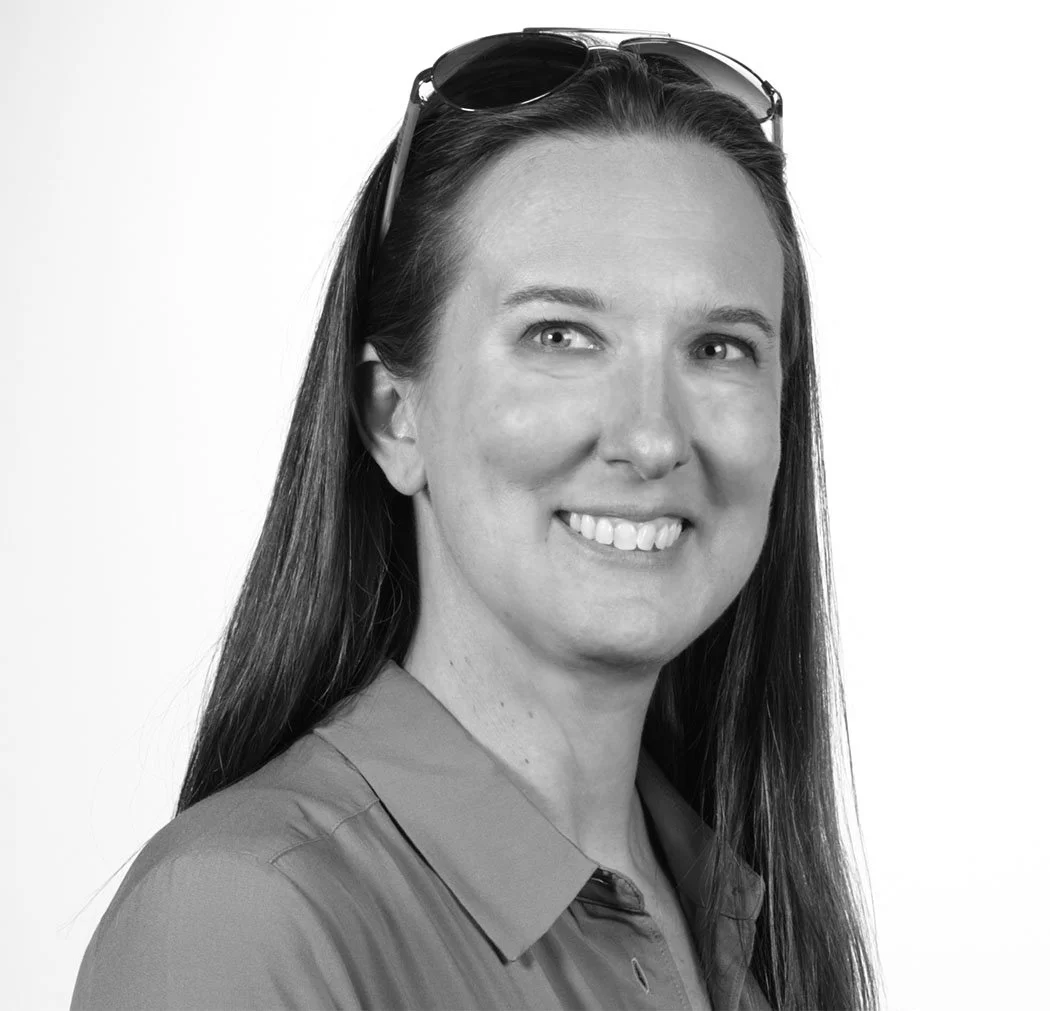 Black and white portrait of a smiling woman named Brett Cambria with long hair and sunglasses on her head, wearing a collared shirt.