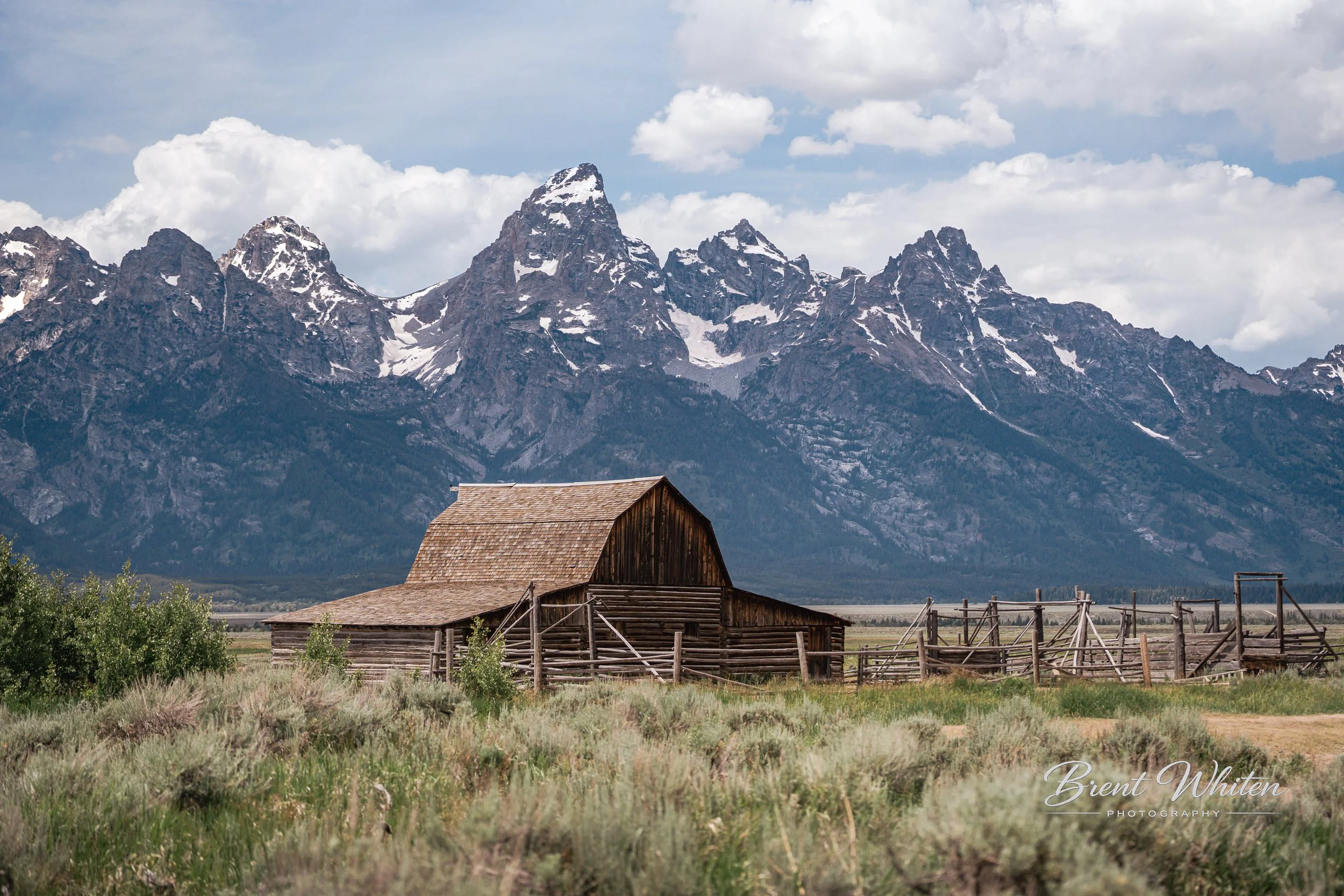 A rustic wooden barn with a weathered roof sits in front of a mountain range with snow-capped peaks. The sky is partly cloudy, and the scene is surrounded by green grass and a wooden fence.