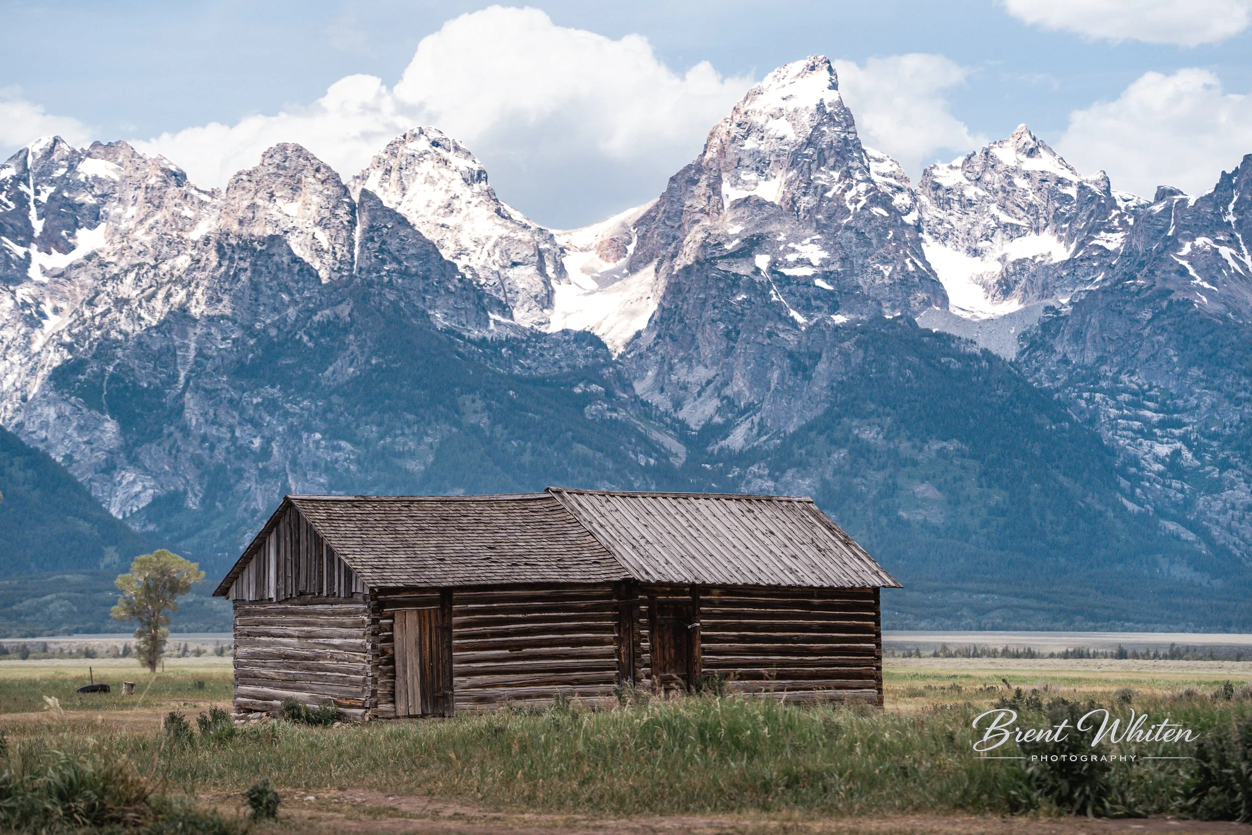 A rustic wooden barn in the foreground with a mountain range covered in snow behind it and partly cloudy skies.