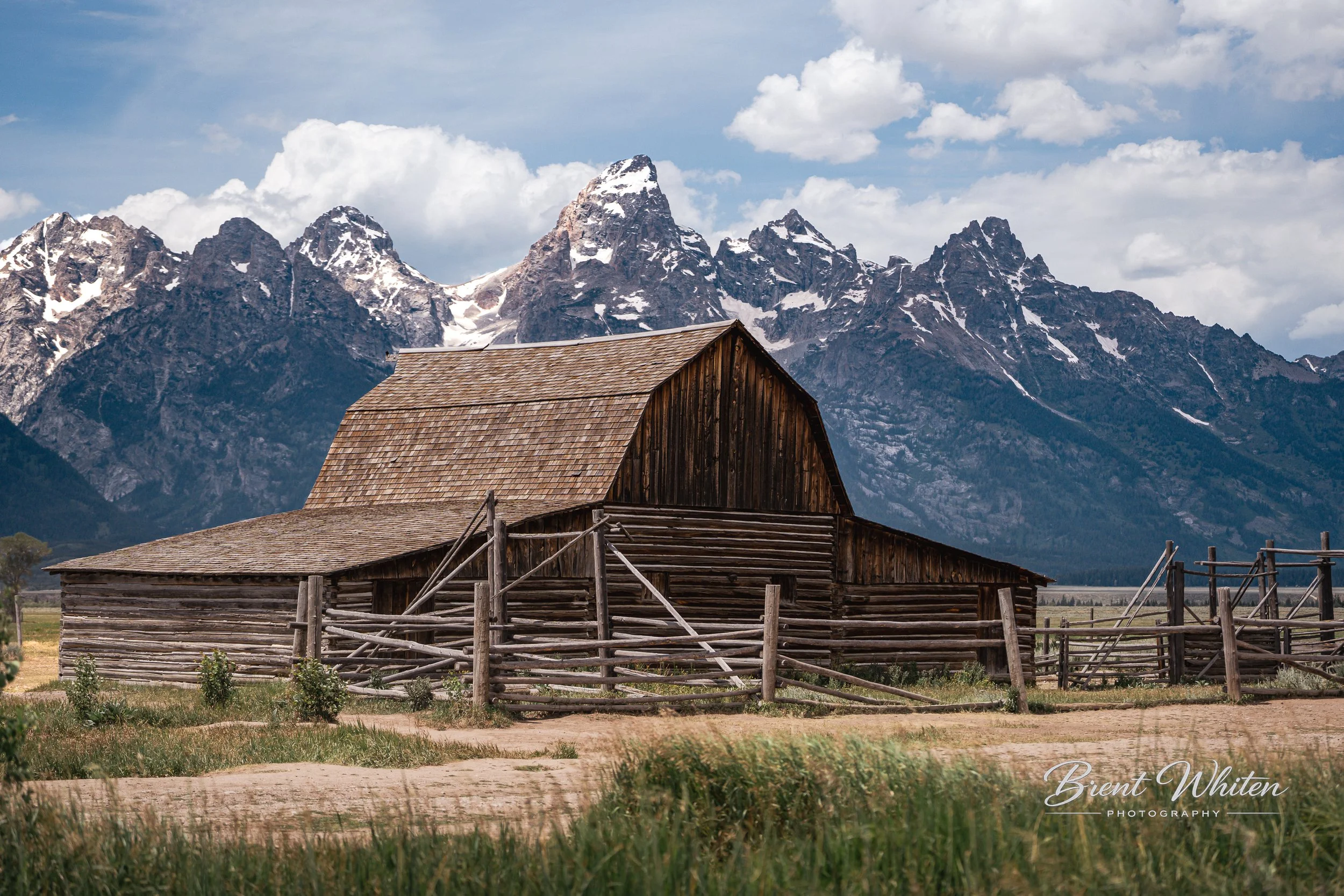 A rustic wooden barn with a sloped roof and fencing in front, set against a backdrop of snow-capped mountains and a partly cloudy sky.