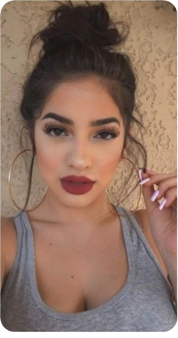 Woman with hoop earrings and red lipstick takes a selfie against a textured wall.
