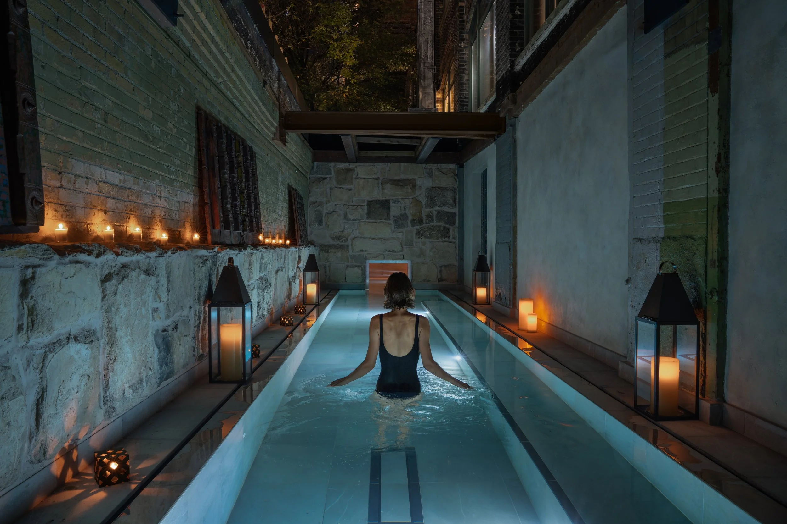 A woman in a black swimsuit standing in a narrow indoor pool, surrounded by lit candles in lanterns, with candles on a stone wall to the left, and soft lighting creating a relaxing ambiance.