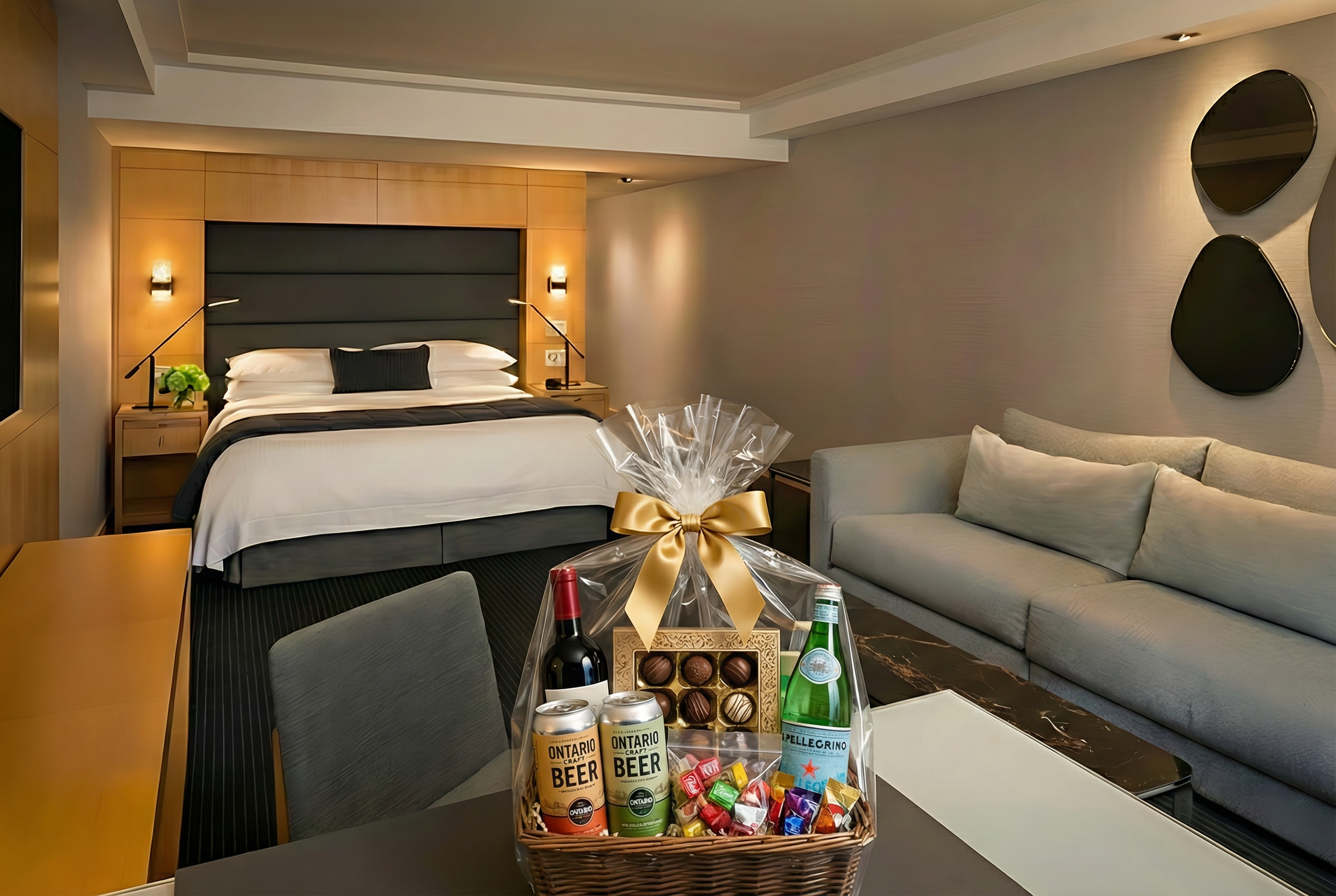 A hotel room with a king-sized bed, wall-mounted lights, a gray headboard, a beige sofa, and wall art. On the table, there is a gift basket with chocolates, a box of assorted chocolates, a bottle of sparkling water, a bottle of wine, and cans of craft beer.