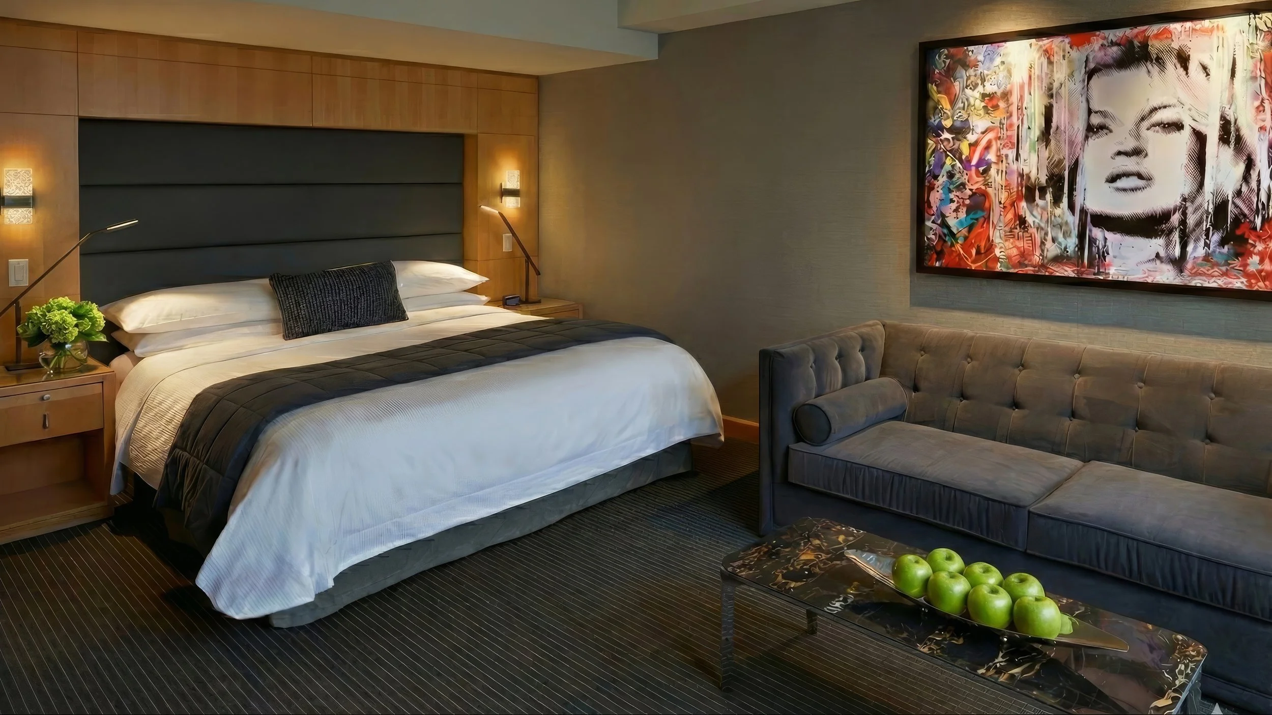 A modern hotel room with a king-size bed, dark gray headboard, and white linens, next to a gray velvet sofa and a black coffee table with green apples. A colorful abstract portrait of Marilyn Monroe hangs on the wall.
