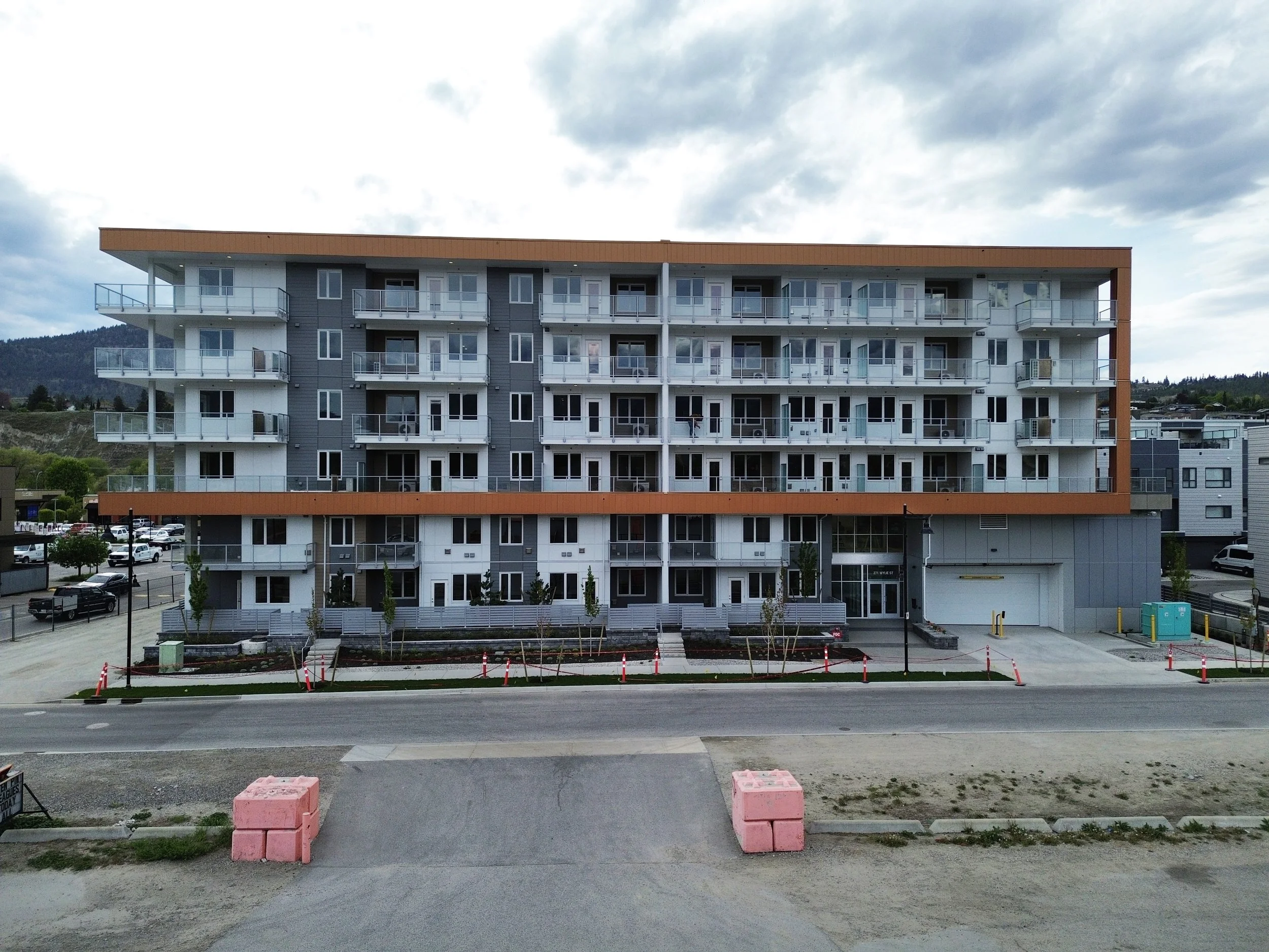 Modern multi-story apartment building under construction with orange accents, white balconies, and glass railings, with construction barriers and vehicles in the foreground, and a cloudy sky above.